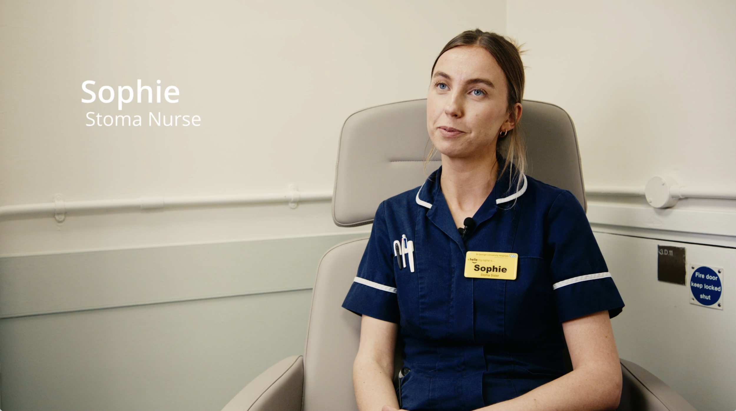 A nurse named Sophie in a navy blue uniform with a name badge sitting in a hospital room.