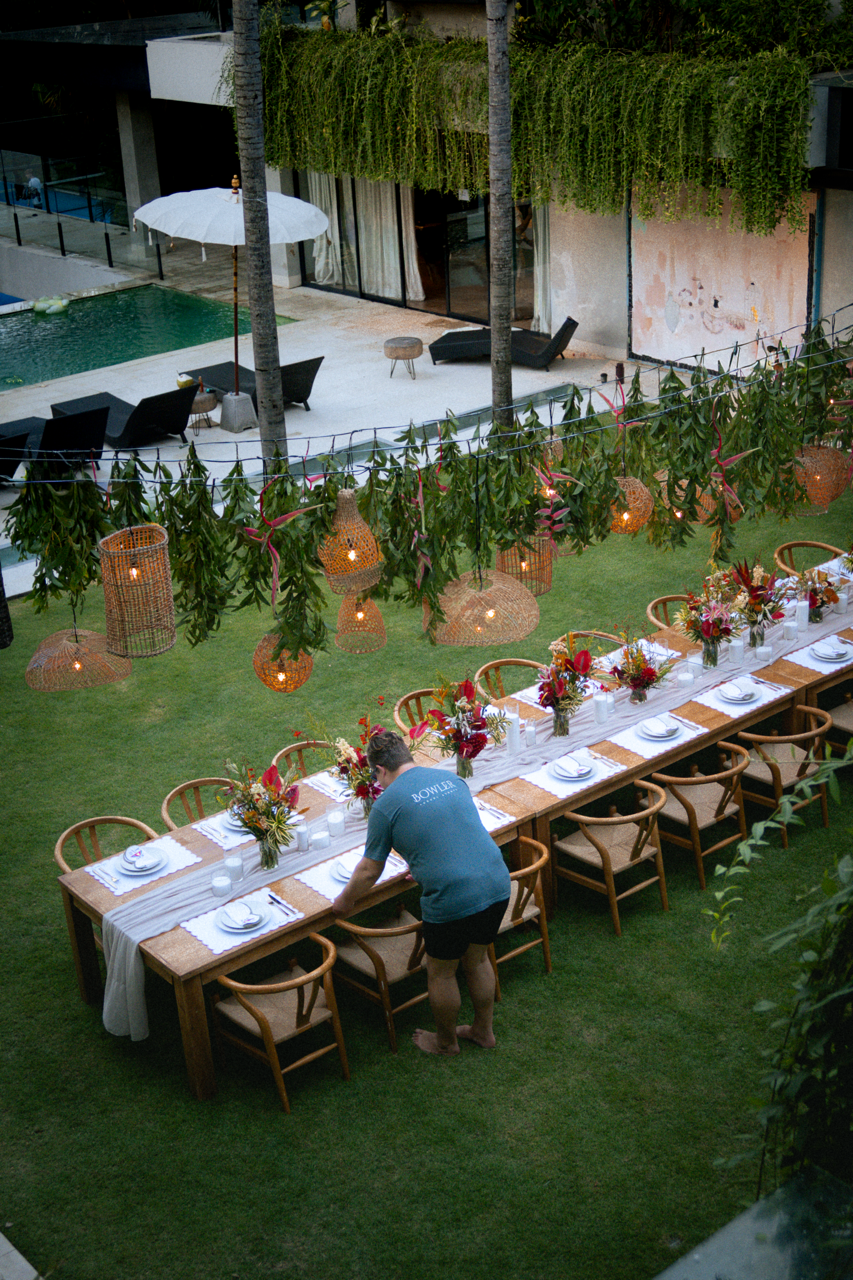 A long outdoor dining table set with white napkins, plates, and colorful floral centerpieces under hanging wicker lanterns and greenery in a backyard garden.