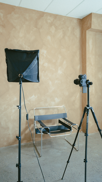 Photography setup with a softbox light on a stand, a black chair, and a camera on a tripod in a studio with beige walls.