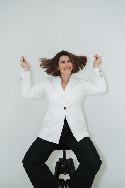 Woman sitting on a stool with arms raised, smiling, in a white blazer and black pants against a plain white background.