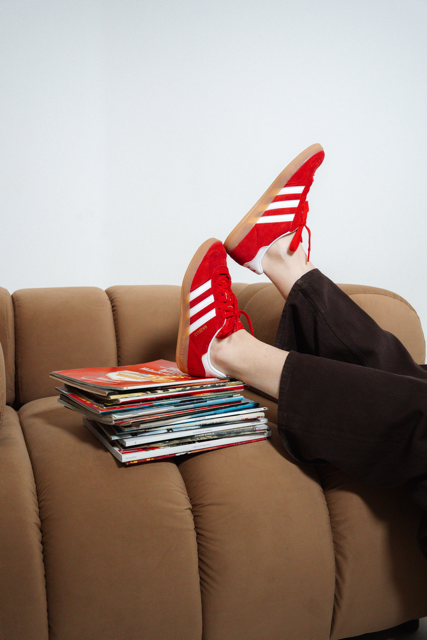 Person wearing red sneakers with white stripes resting their feet on a stack of magazines on a tan sofa