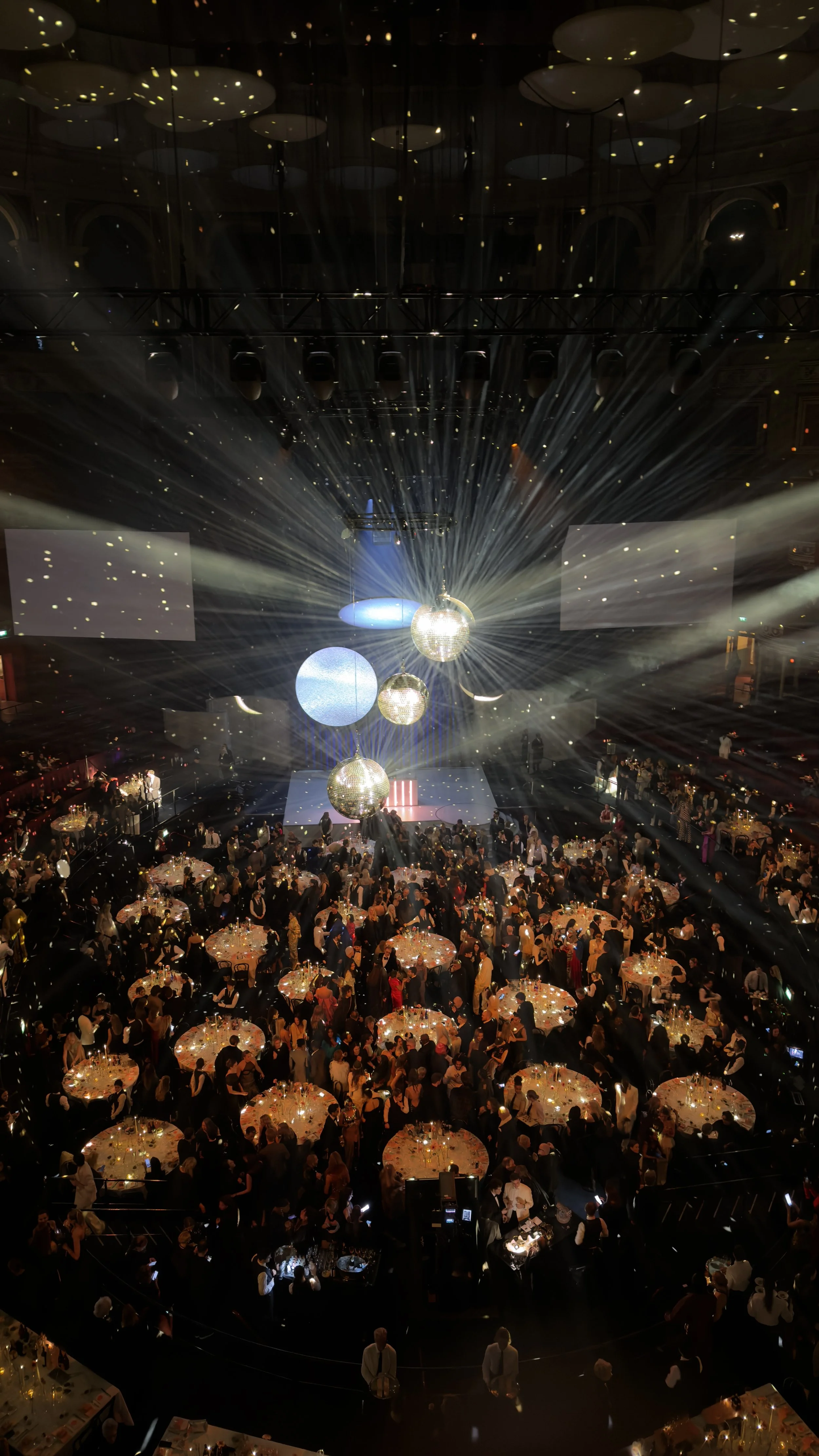 A large banquet hall filled with round tables of people attending a formal event, with a stage in the center decorated with large disco ball ornaments and bright lights.