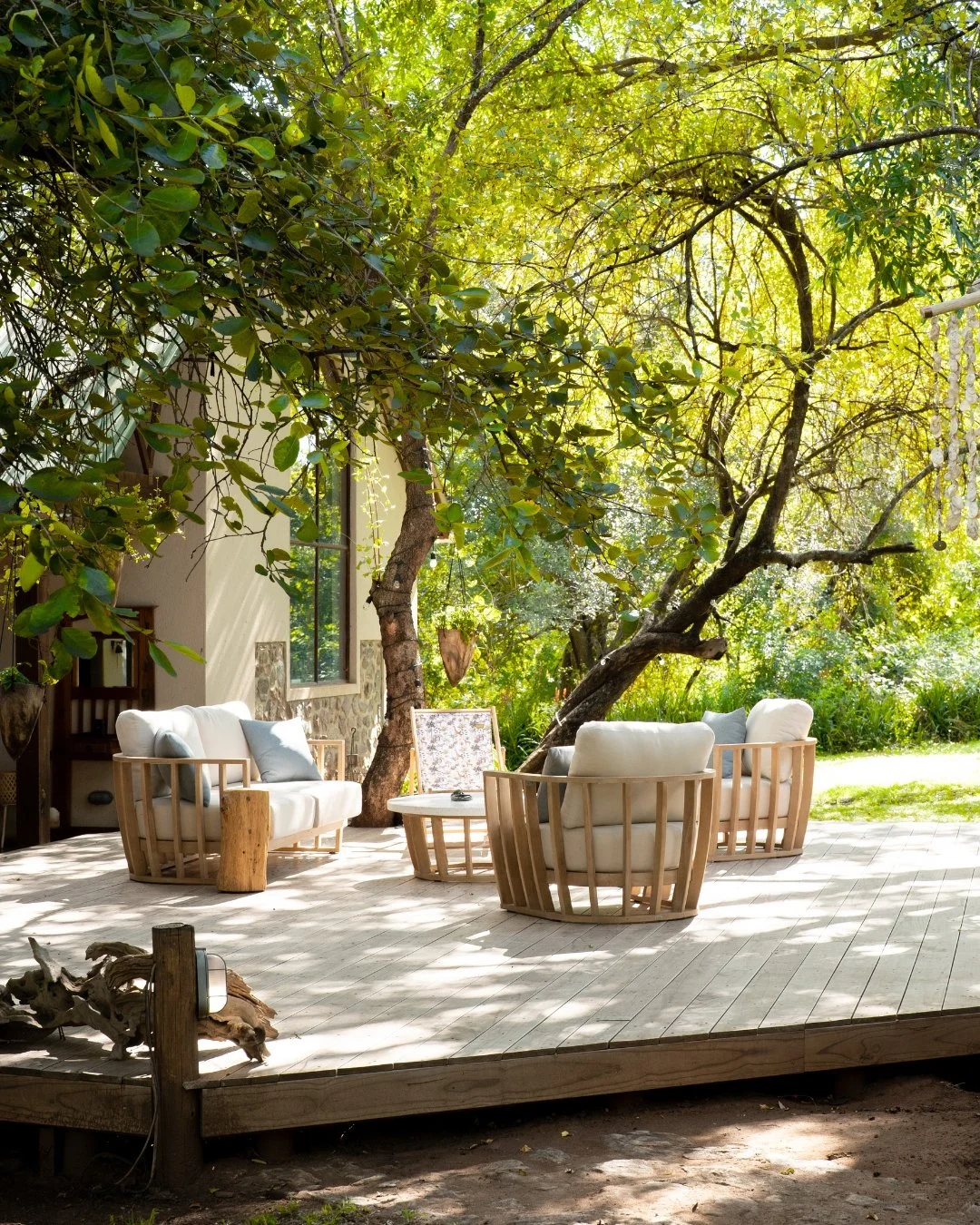 A wooden deck with outdoor furniture, including white cushioned chairs and a small table, surrounded by lush green trees and foliage in sunlight.