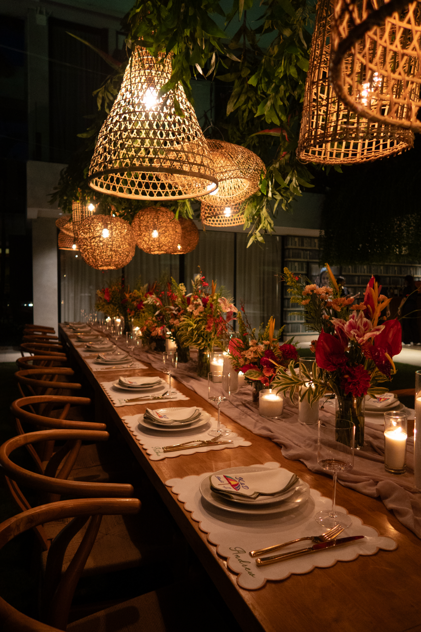 A long dining table set for a celebration with plates, glasses, and silverware, decorated with multiple vases of colorful flowers and flickering candles. Above, there are woven pendant lights hanging from the ceiling, creating warm ambient lighting in a cozy indoor space.