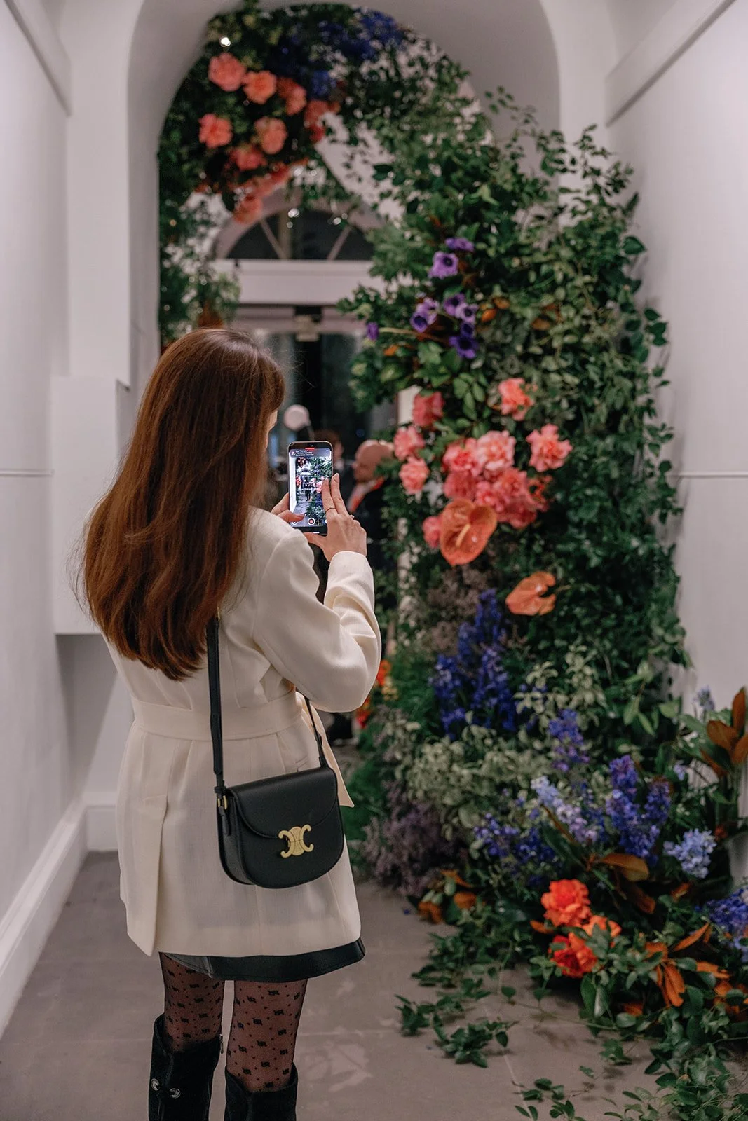 A woman with long brown hair takes a photo of a floral arrangement in a white-walled indoor space. The arrangement features pink, purple, and blue flowers with green foliage.