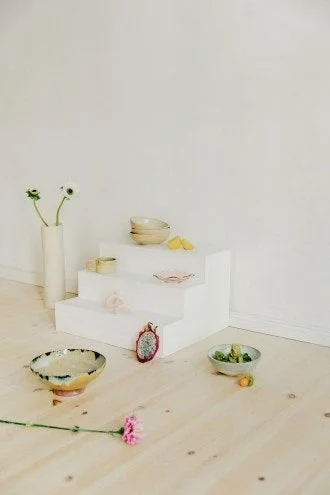 Minimalist display with white stairs, bowls, and flowers on a light wooden floor against a white wall.