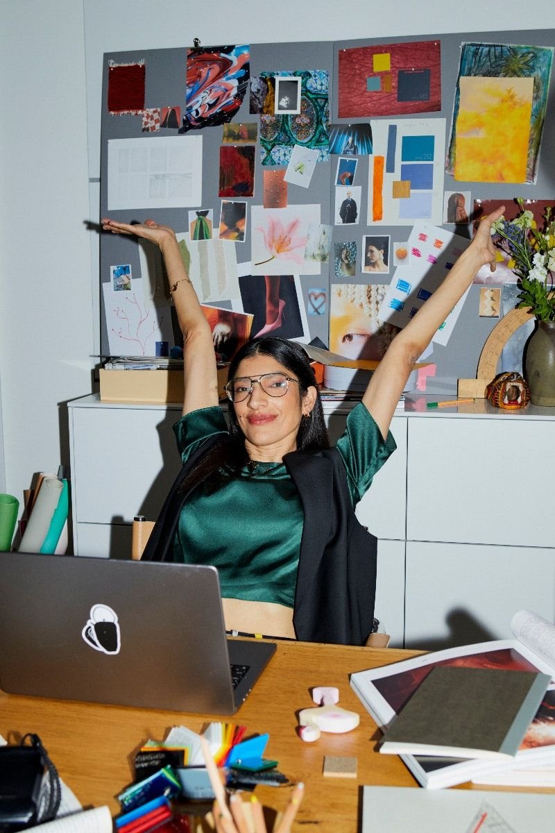Woman with glasses smiling at camera, stretching arms, sitting at a desk with creative artwork and photos on a gray board behind her.