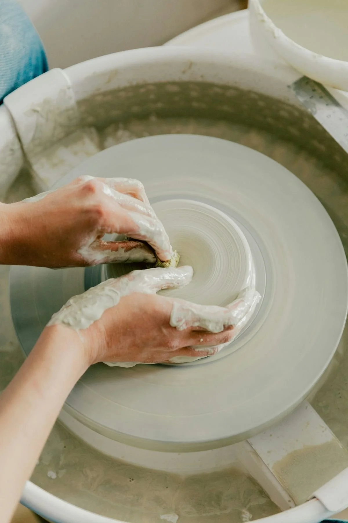 A person is shaping clay on a pottery wheel with both hands covered in clay.