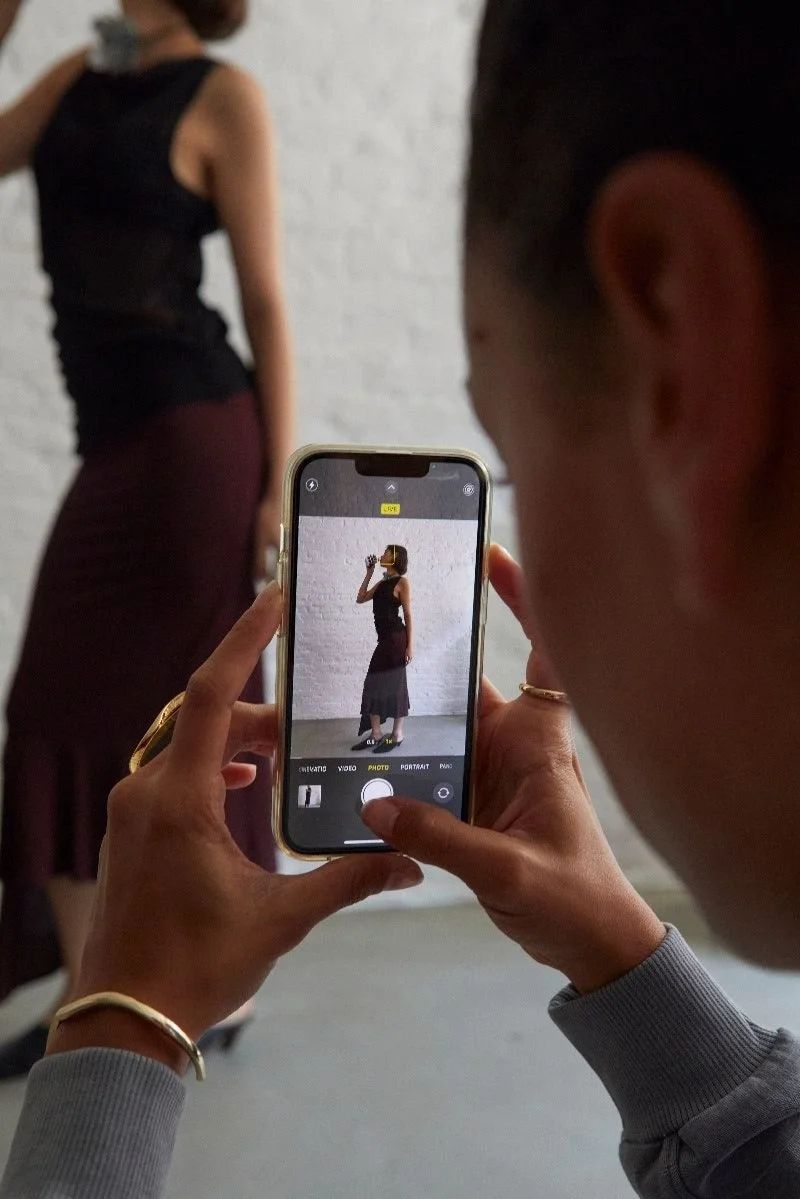 A person taking a photo of a woman standing against a white brick wall, with the phone's screen showing her pose.