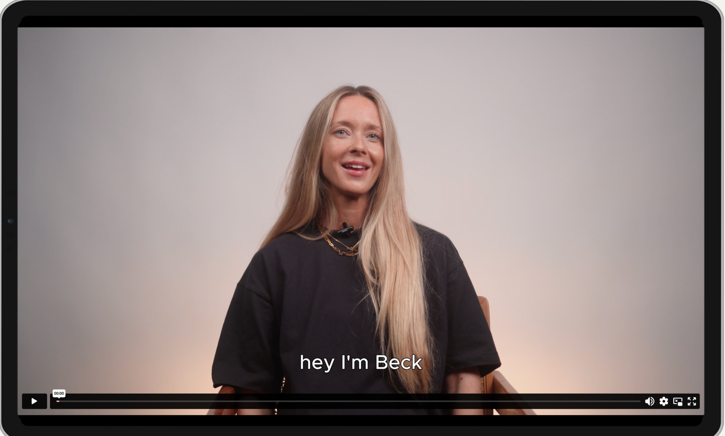 A woman with long blonde hair, wearing a black shirt and a gold chain necklace, sitting on a chair against a plain light gray background, smiling at the camera with subtitles that say "hey I'm Beck".