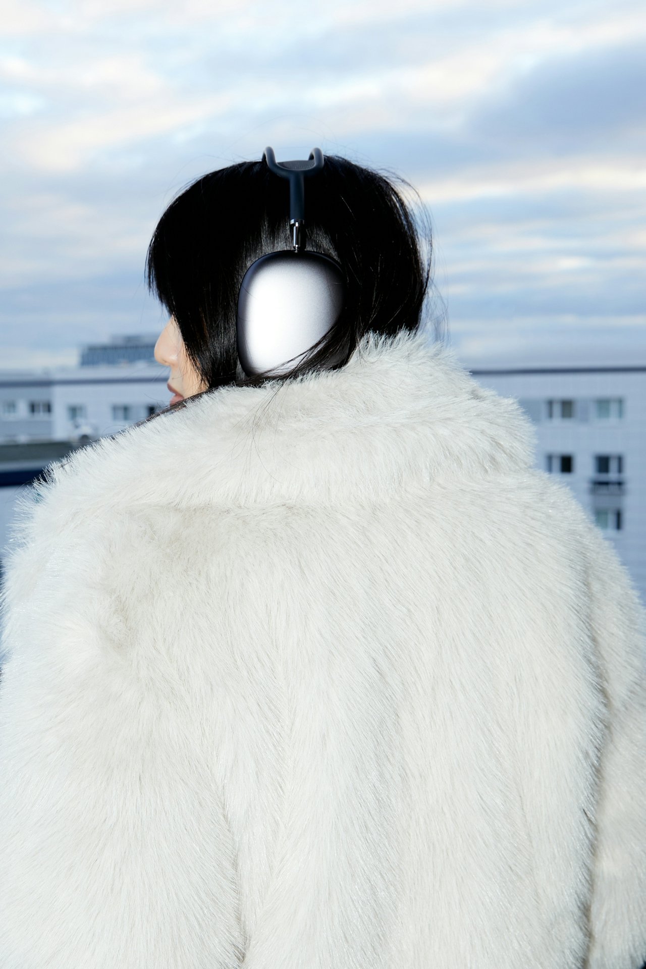A woman with black hair wearing a white fur coat and large over-ear headphones standing outdoors on a cloudy day.