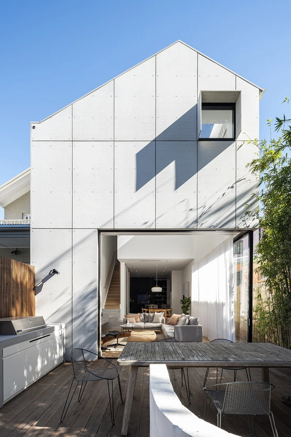 Modern two-story house with large open patio and outdoor dining area, view into living room with sofa and kitchen, blue sky, and greenery on the side.