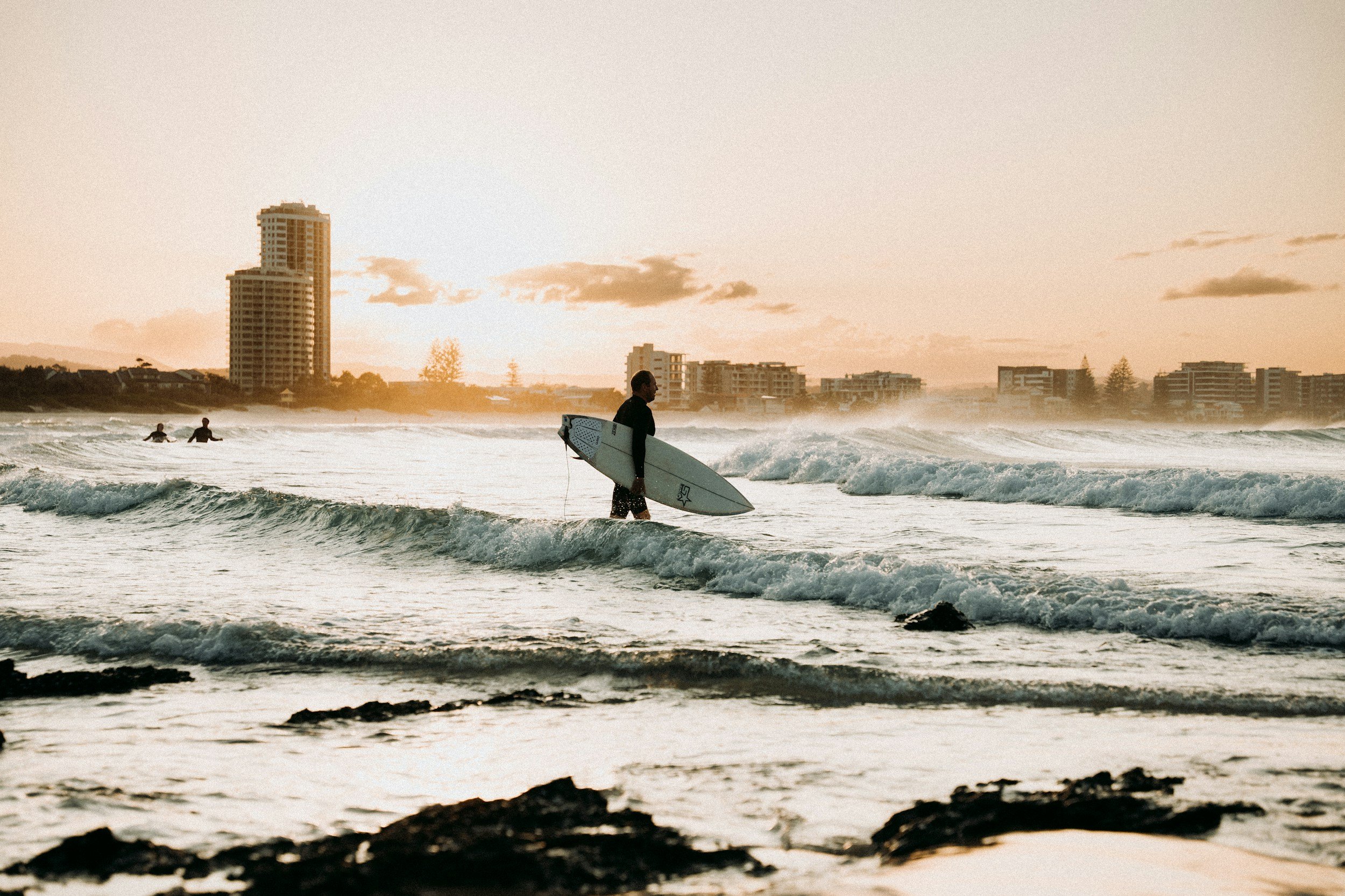 A person standing in the ocean holding a surfboard at sunset, with a city skyline in the background.