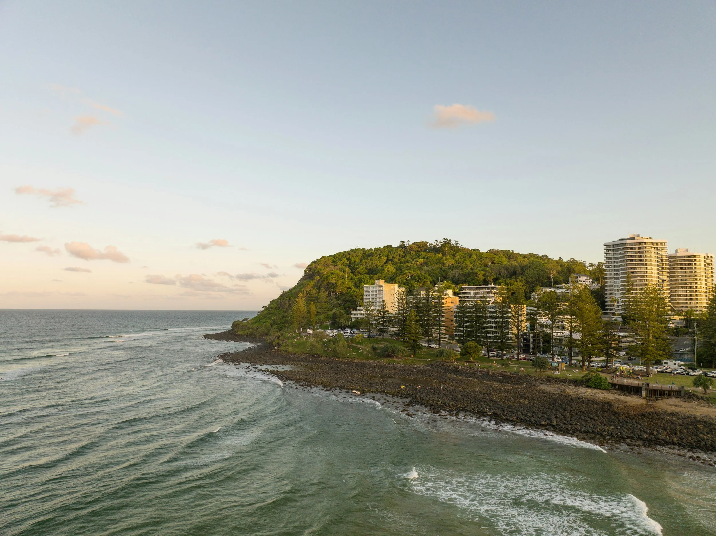 Coastal view of a hillside with residential buildings and trees, next to the ocean with gentle waves, under a partly cloudy sky during sunset.