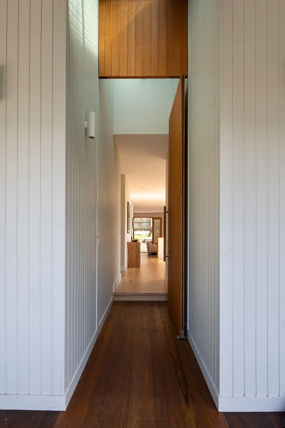 A narrow hallway with wood-paneled and white shiplap walls, leading to a living room with large windows and outdoor view.
