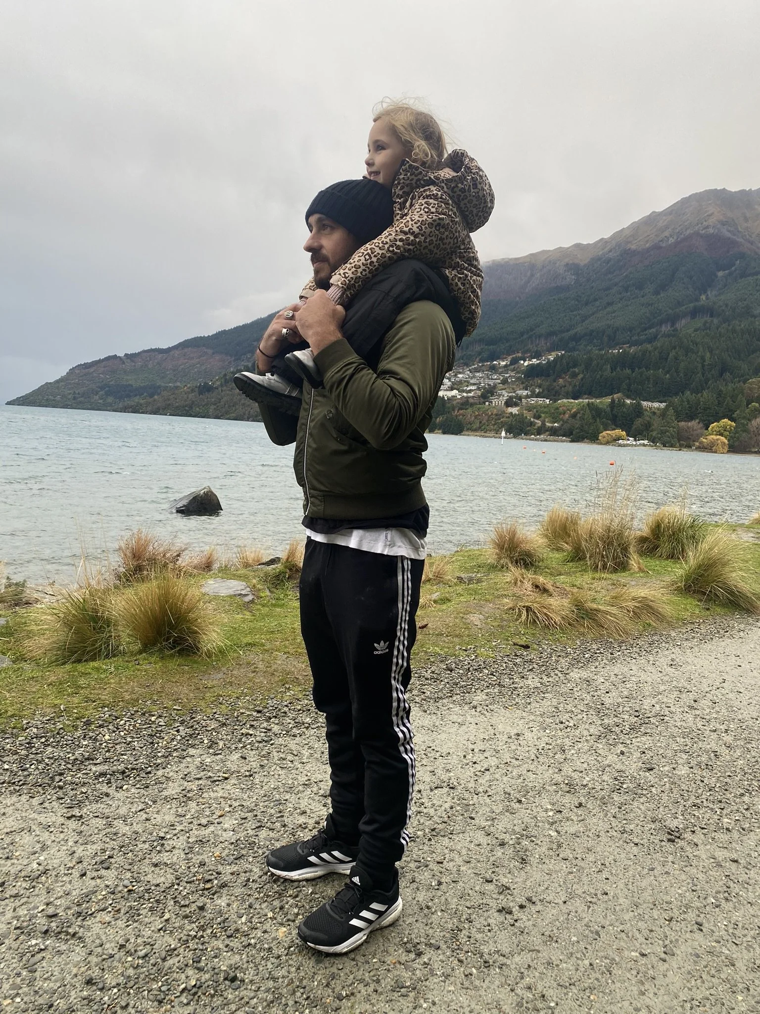 A man carries a young girl on his shoulders by a lakeshore with mountains and trees in the background, overcast sky.