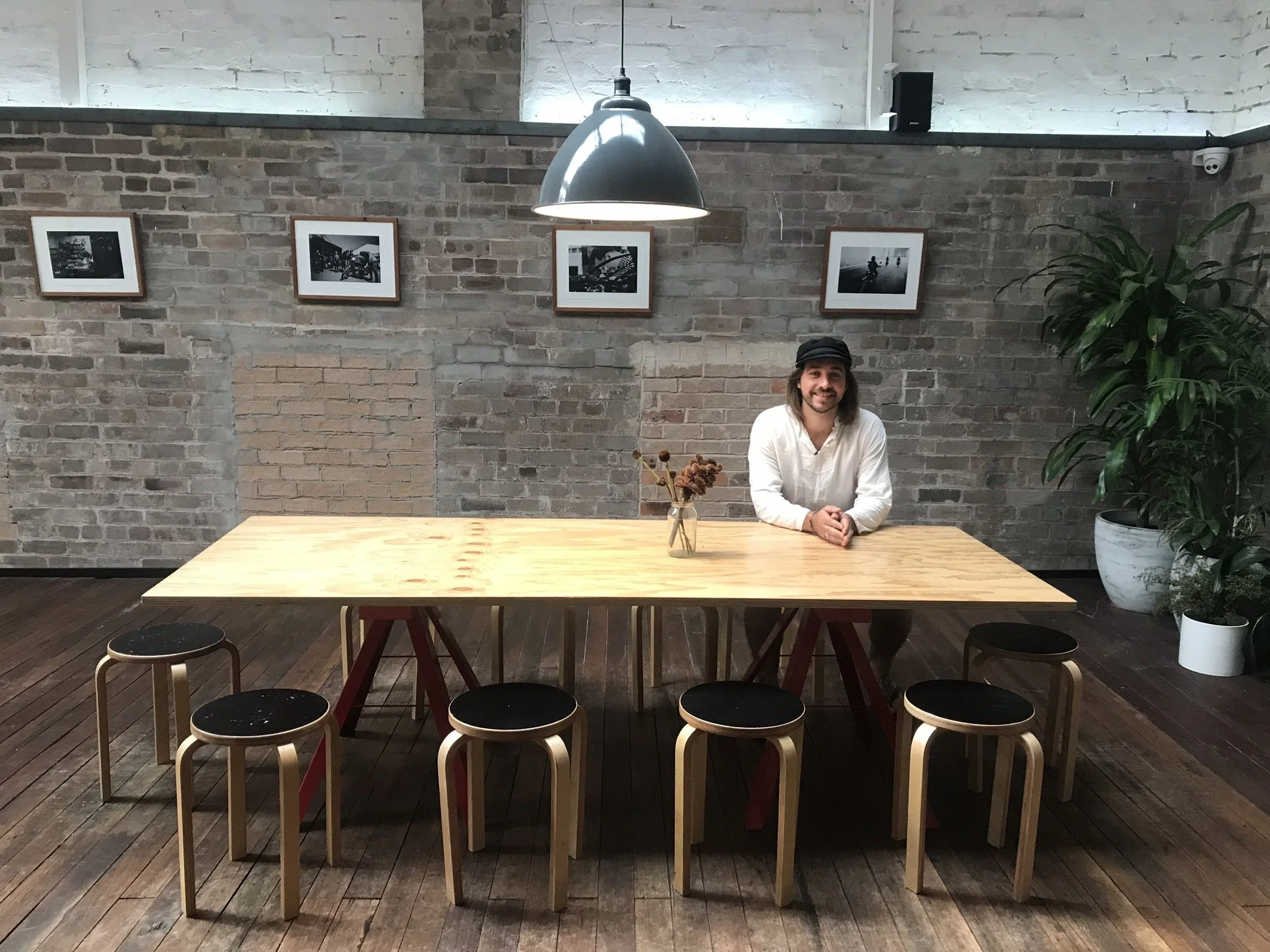 A man with long hair and a beard, wearing a white shirt and a black cap, sitting at a large wooden table in a modern, industrial-style room with brick walls, hardwood flooring, and black and white framed photographs on the wall. There is a vase with dried flowers on the table and large plants in the corner.