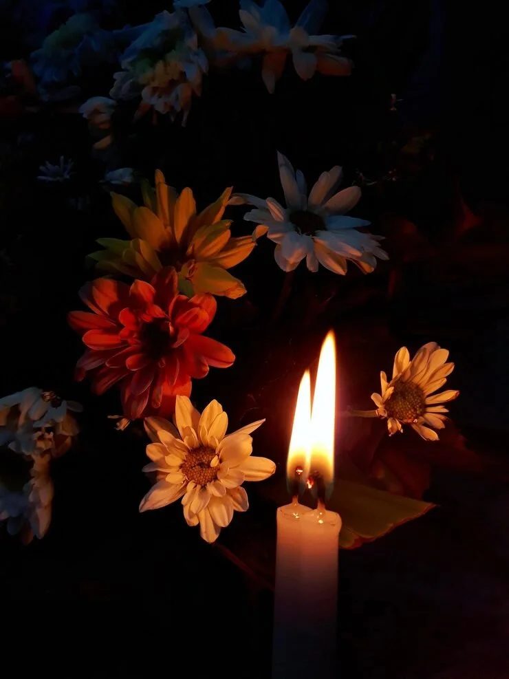 A lit white candle with two flames surrounded by colorful flowers, including daisies and other blooms, in a dark setting.
