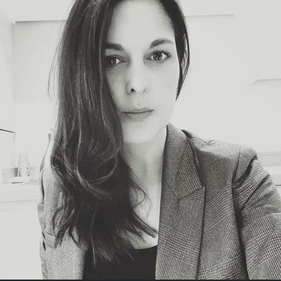 A woman with shoulder-length, wavy dark hair taking a selfie in a black and white photo. She is wearing a plaid blazer over a dark top, with a neutral expression. In the background, a room with a tray holding glasses and bottles is visible.