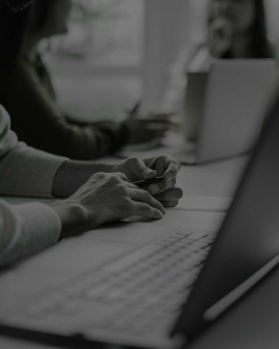 People working at a desk with laptops, using smartphones, in a dimly lit room.