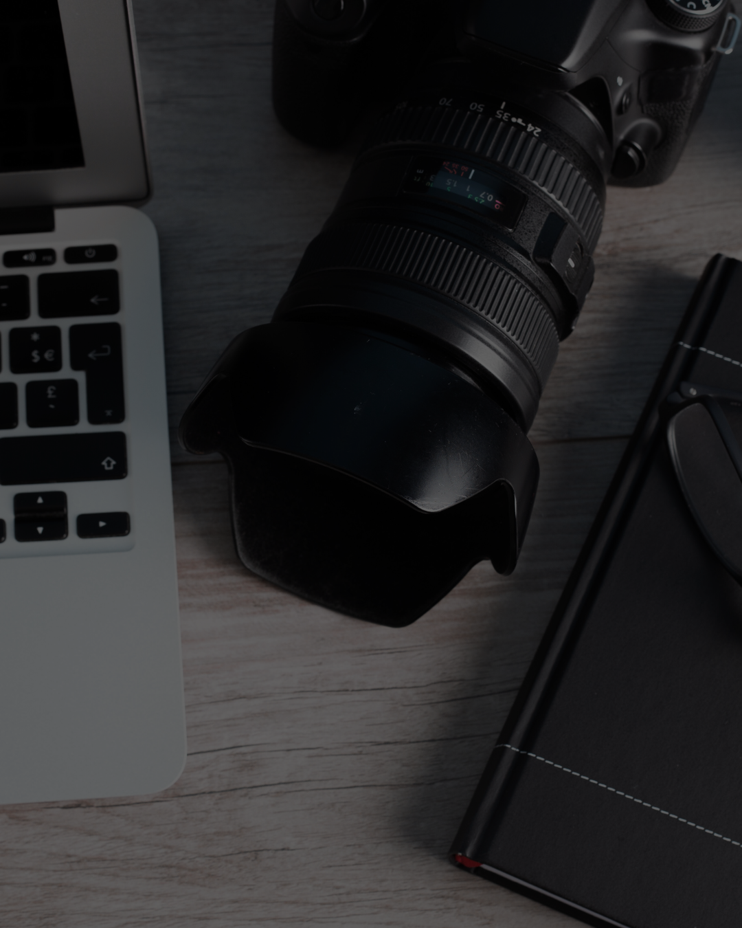 A DSLR camera with a zoom lens, a laptop keyboard, a black notebook, and a smartphone on a wooden desk.