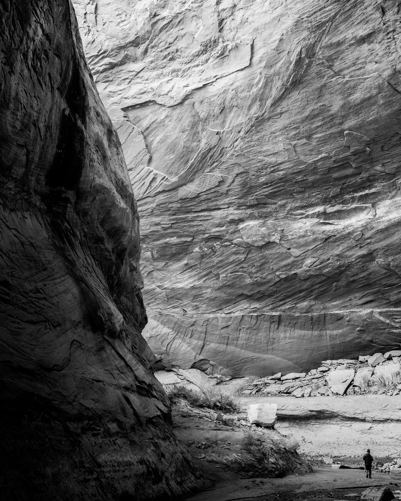 Black and white photograph of a narrow canyon with towering rock walls. A small figure of a person is walking along the rocky, sandy ground near the bottom right corner, emphasizing the scale of the canyon.