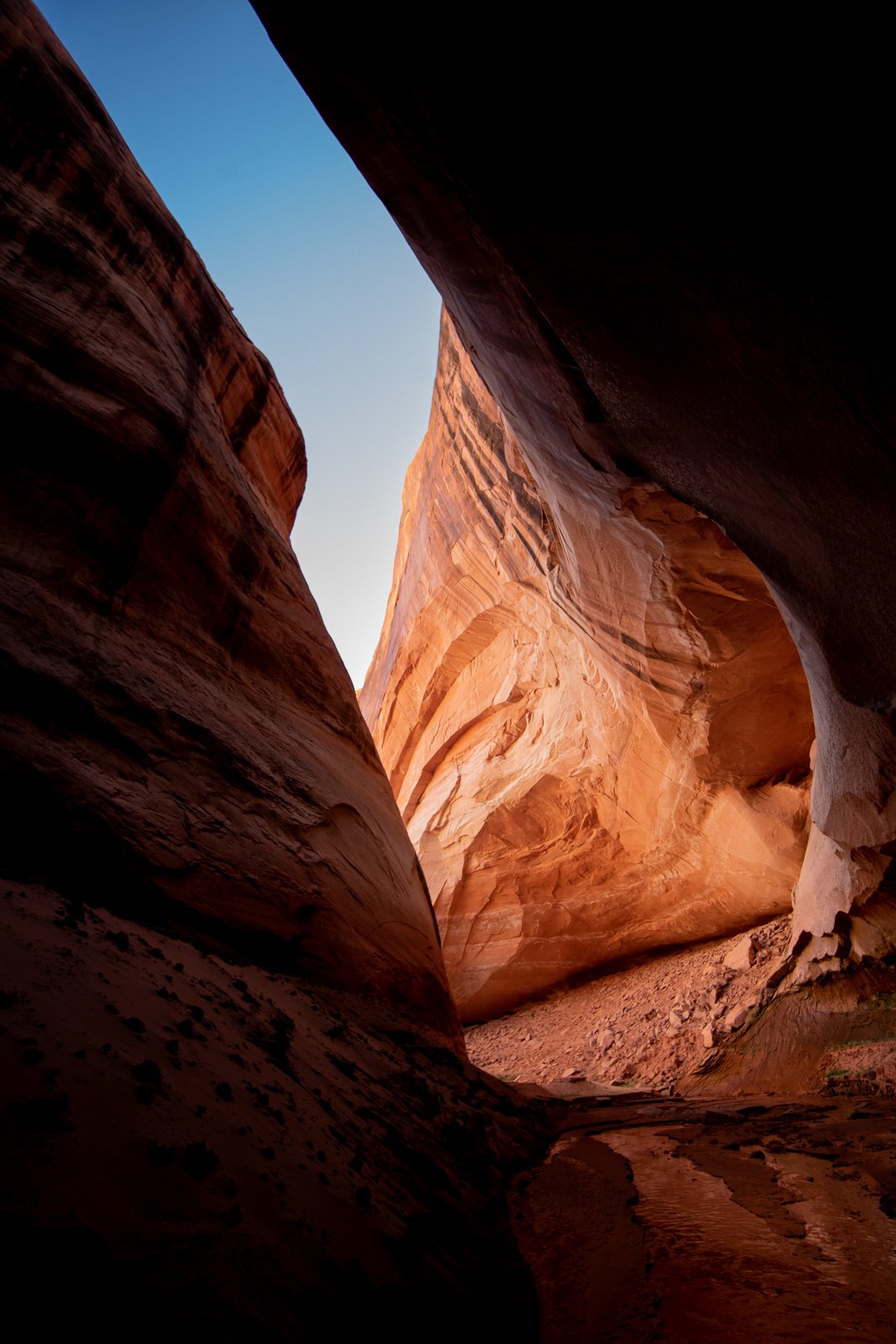 Narrow slot canyon with smooth, layered red and orange sandstone walls and a bright blue sky visible above.