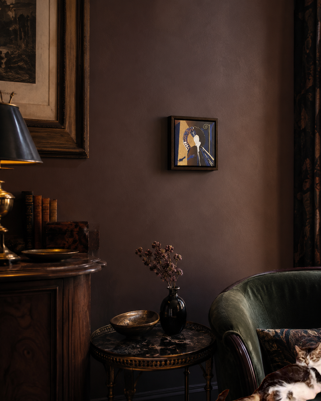 A cozy room corner with a round marble table holding a black vase with dried flowers and a brass bowl, a green upholstered armchair partially visible, a dark brown wall with two framed artworks, and warm ambient lighting.