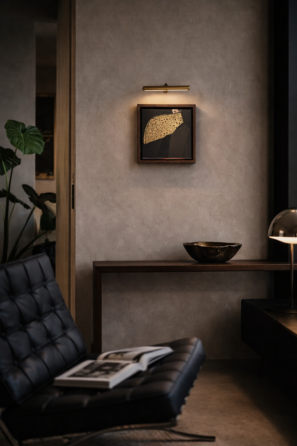 A modern interior room with beige textured walls, a framed abstract artwork illuminated by a small light, a dark decorative bowl on a wooden console, and a black leather tufted lounge chair with an open magazine. A large plant with broad green leaves