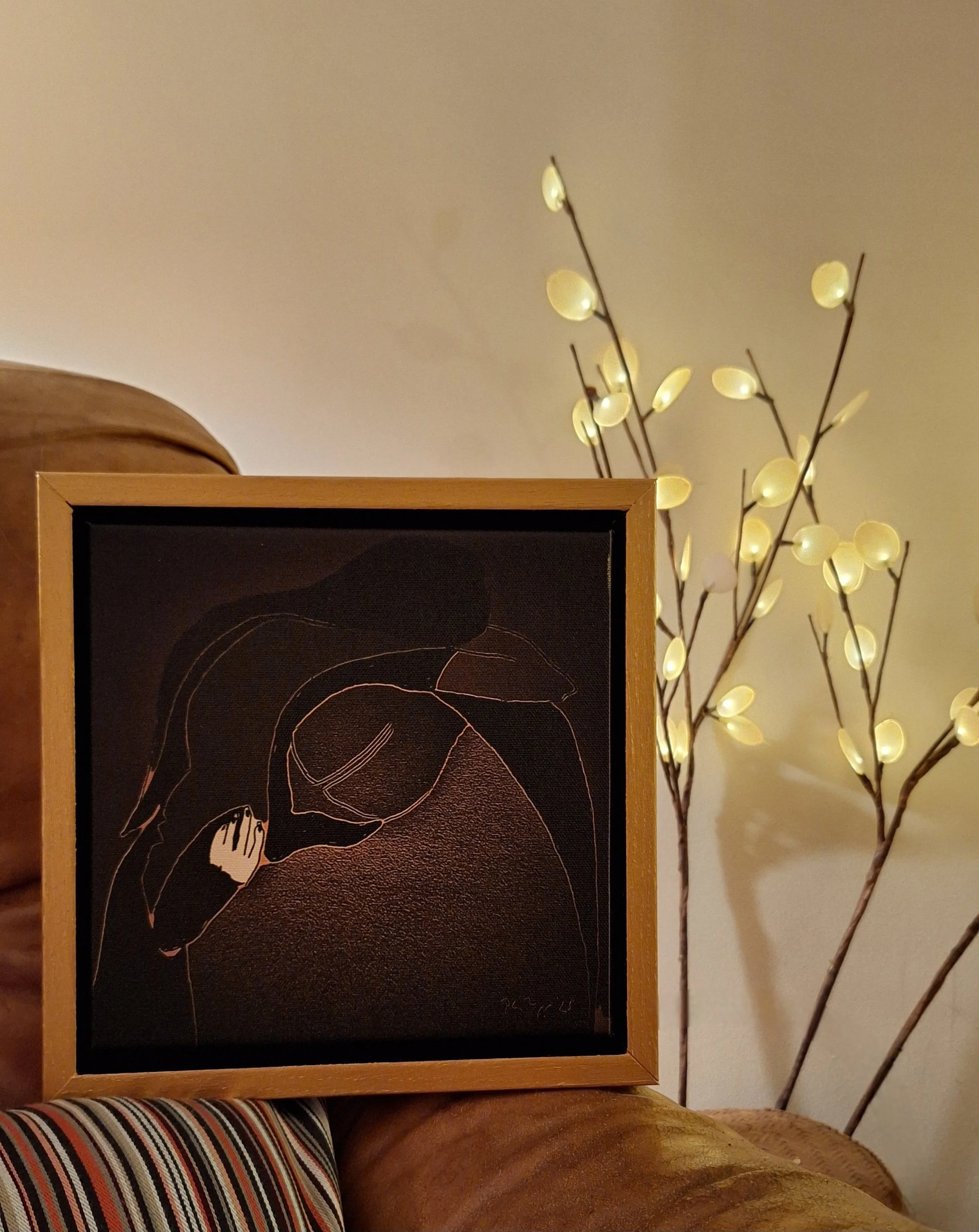 A framed minimalist line art of a woman holding a cup, placed on a sofa with a striped cushion, near a lit decorative branch with small oval lights.