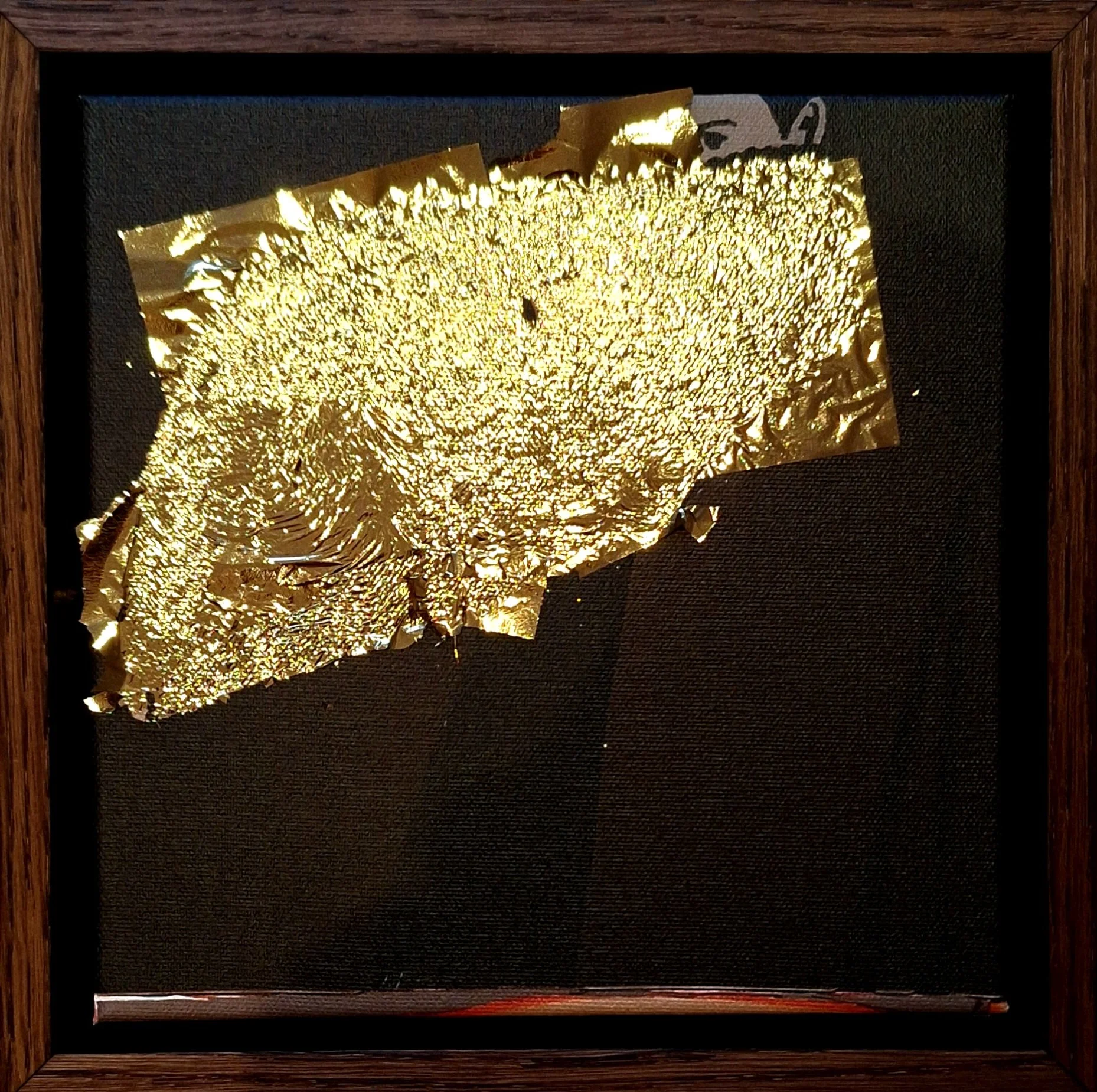 A framed, textured piece of art featuring a gold foil overlay on black background, with some parts crumpled and others smooth, resembling an abstract shape or landmass.