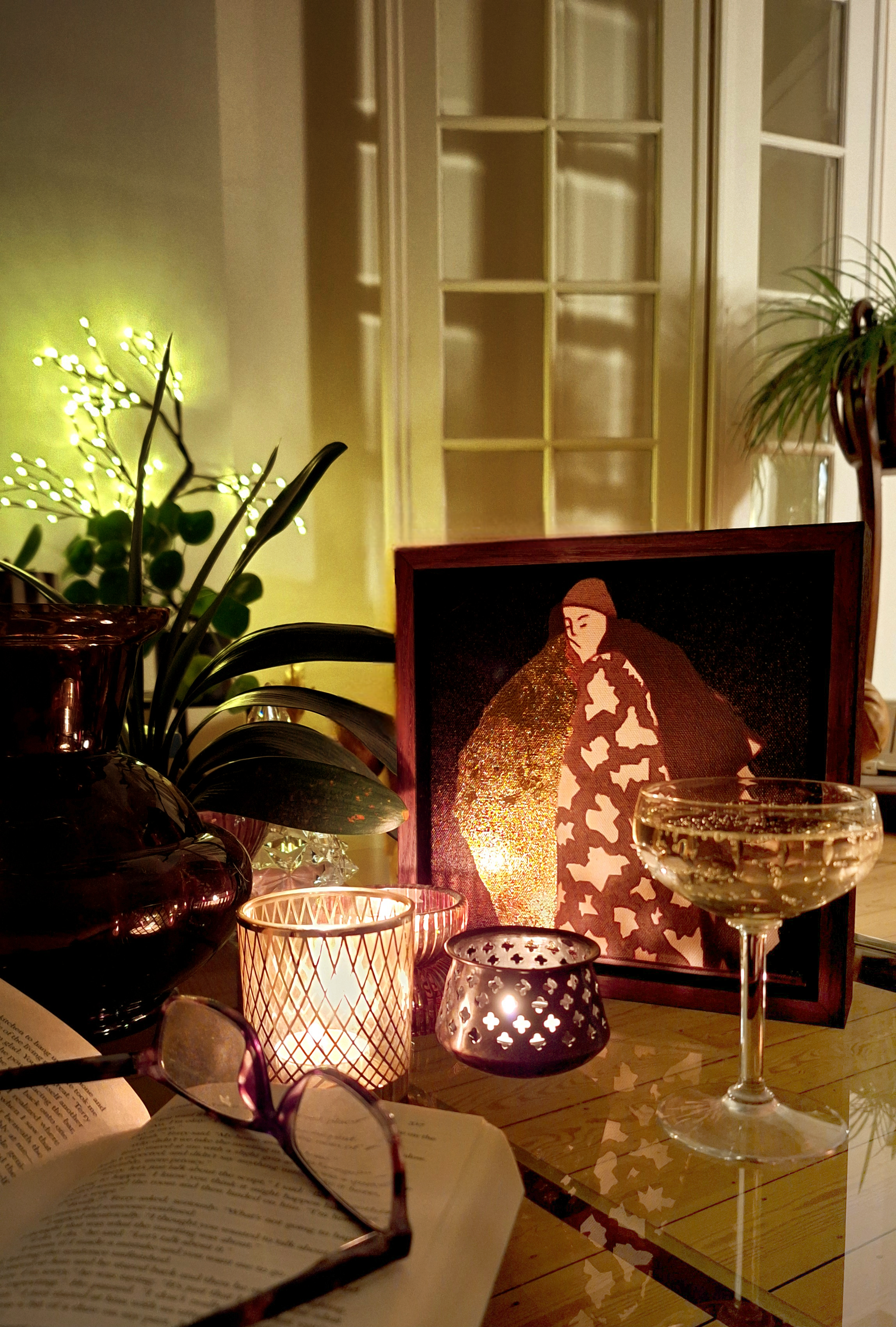 A cozy indoor scene with a table adorned with a lit candle, a glass of white wine, an open book, and a pair of eyeglasses. Behind the table, there is a framed embroidered artwork of a woman in a hat, plants, and decorative candles, creating a warm, i