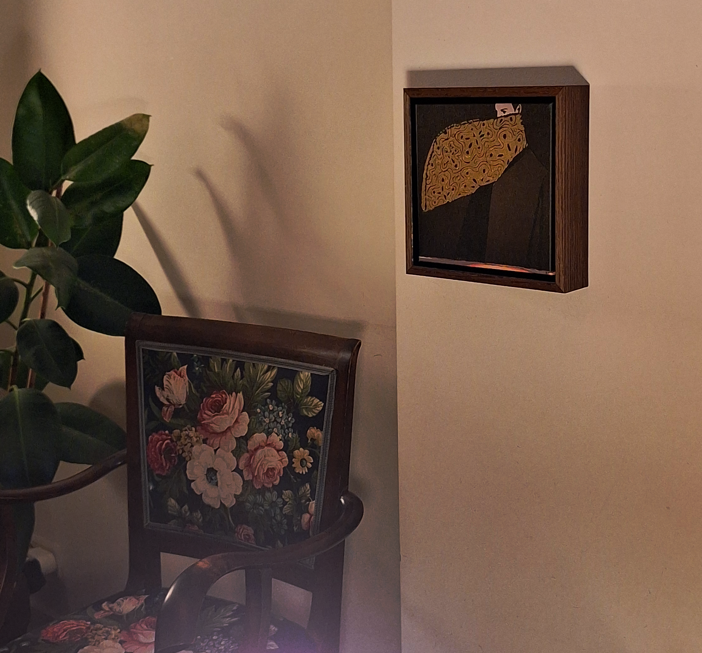 A cozy corner with a wooden chair featuring a floral cushion, a large leafy green houseplant, and two framed artworks on the wall—one depicting a stylized figure with an ornate yellow umbrella, and the other partially visible behind the plant.