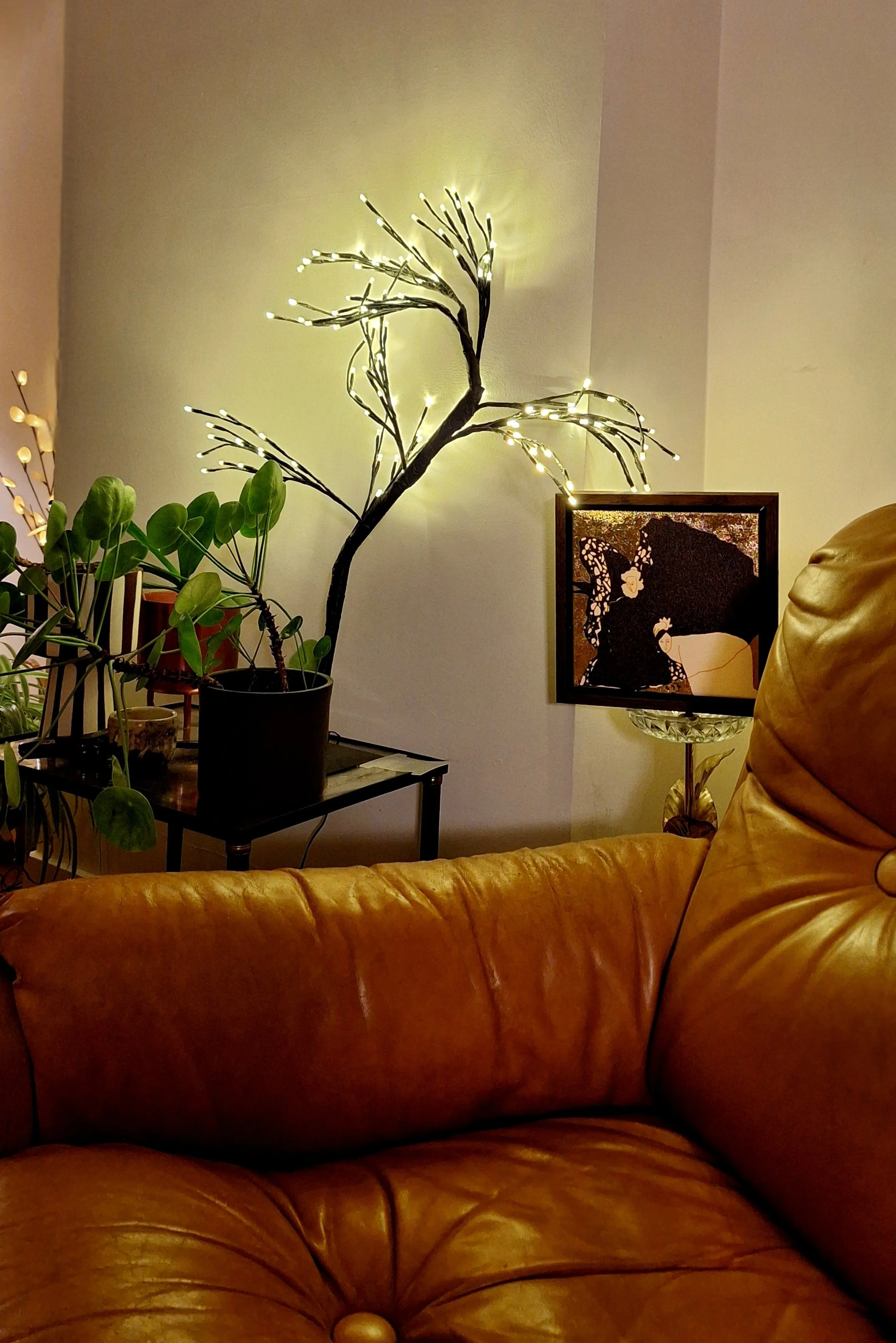 Living room corner with a leather sofa, potted plant with branches and LED lights, a small table with decorative items, and a framed artwork resembling a woman with flowers in her hair.
