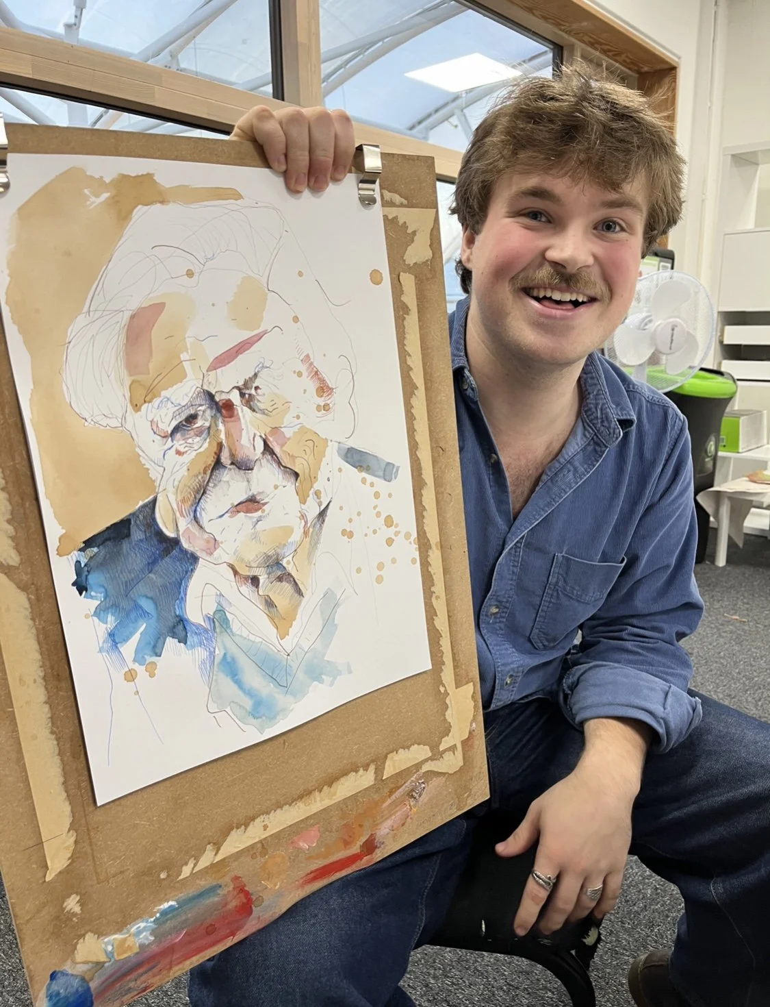 A man with a mustache and blue shirt sitting indoors, smiling and holding up a watercolor portrait of an elderly man in glasses with a bun hairstyle, created on a drawing pad.