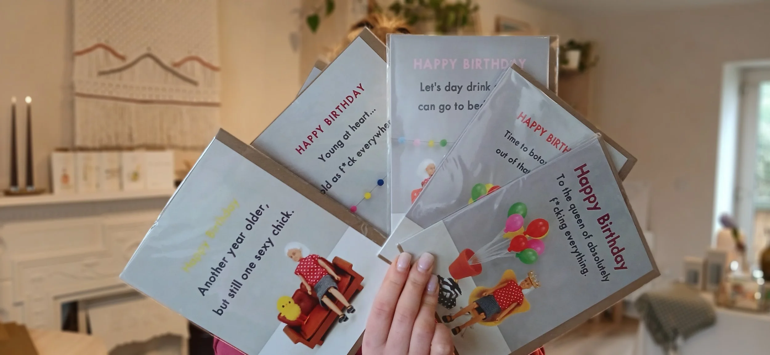 Set of novelty birthday cards with humorous messages and colorful illustrations, held in a person's hand in a cozy indoor setting.