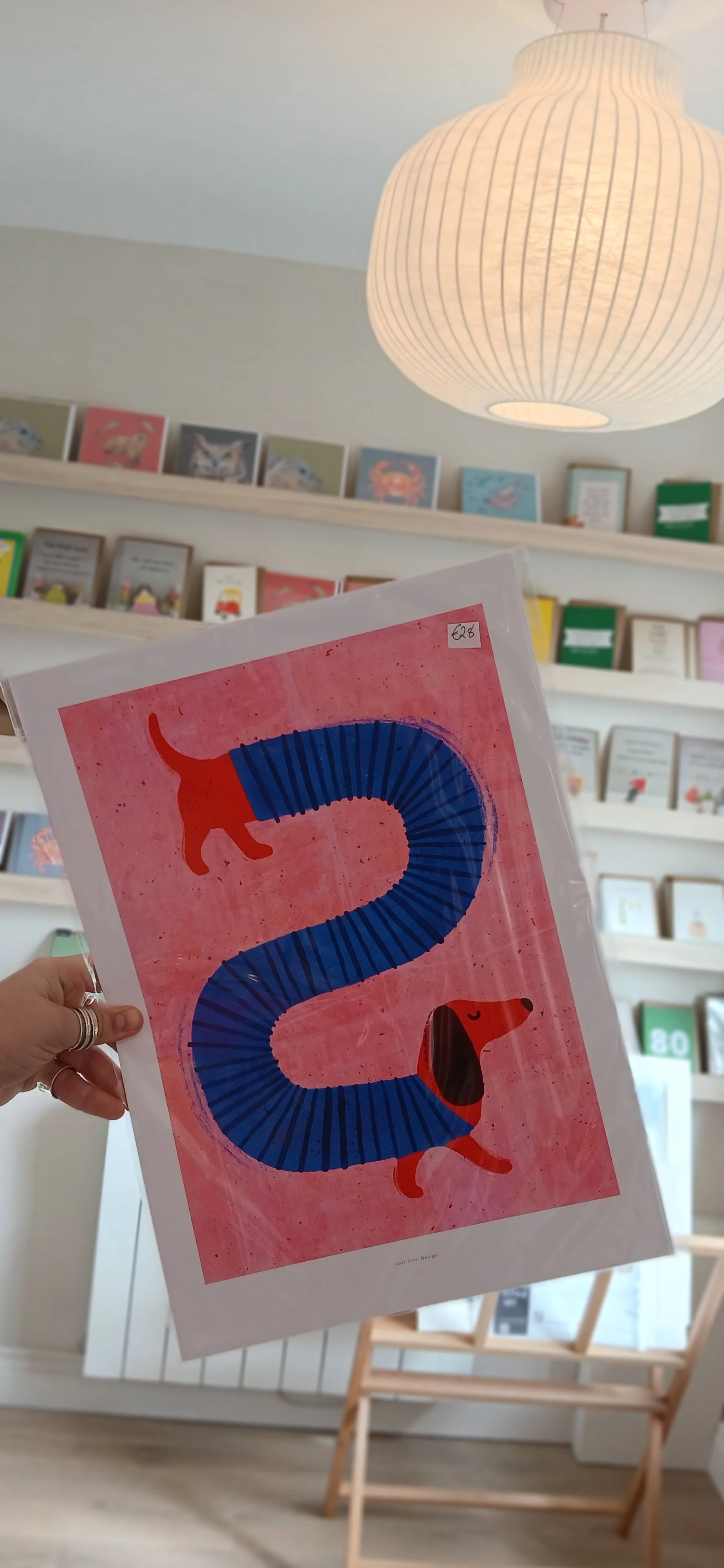 A person holding a colorful illustrated card of a blue snake with a red cat on its tail and head, set against a pink background, in a bookstore with shelves of books in the background and a paper lantern hanging from the ceiling.