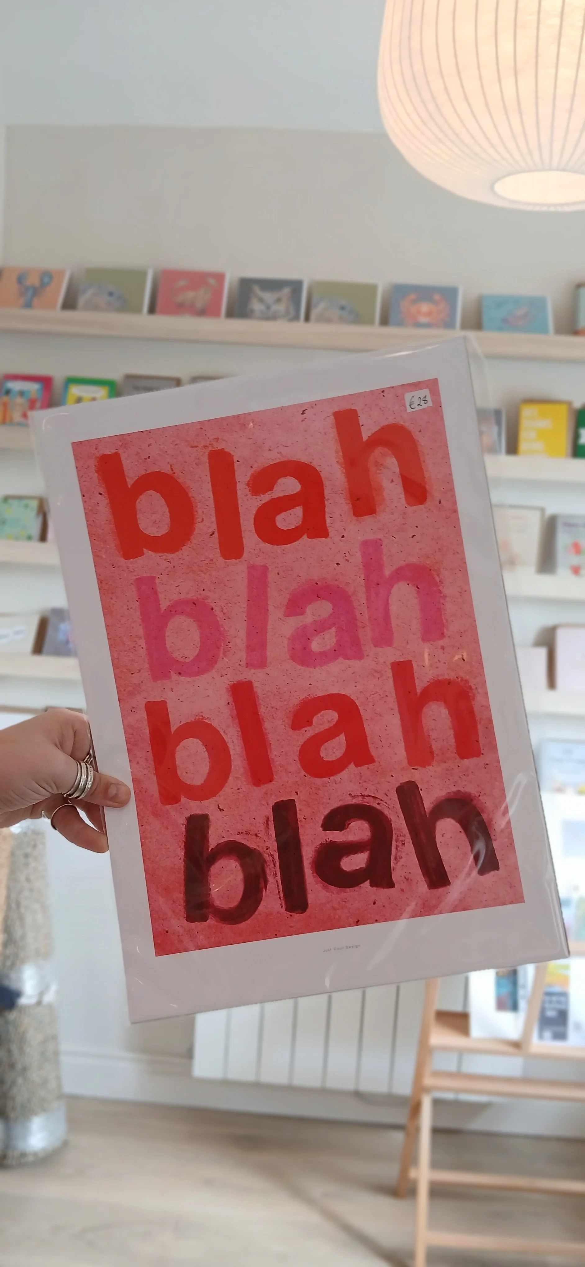 A person holding a pink art print with the word 'blah' repeated four times in different shades, with the bottom last word in black, in a store with shelves of books and artwork in the background.