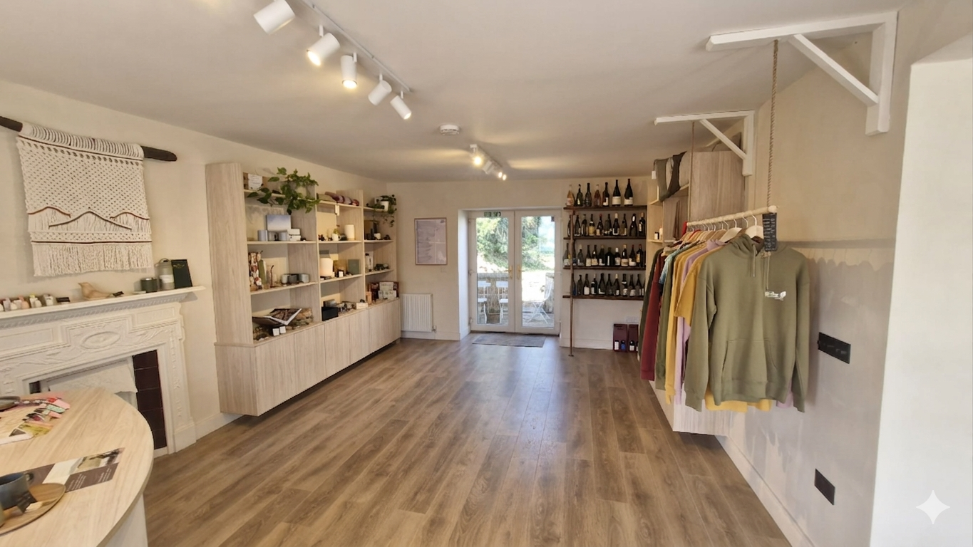 Interior of a boutique shop with wooden flooring, white walls, open shelves displaying various items, a clothing rack with hoodies and shirts, and glass doors leading outside.