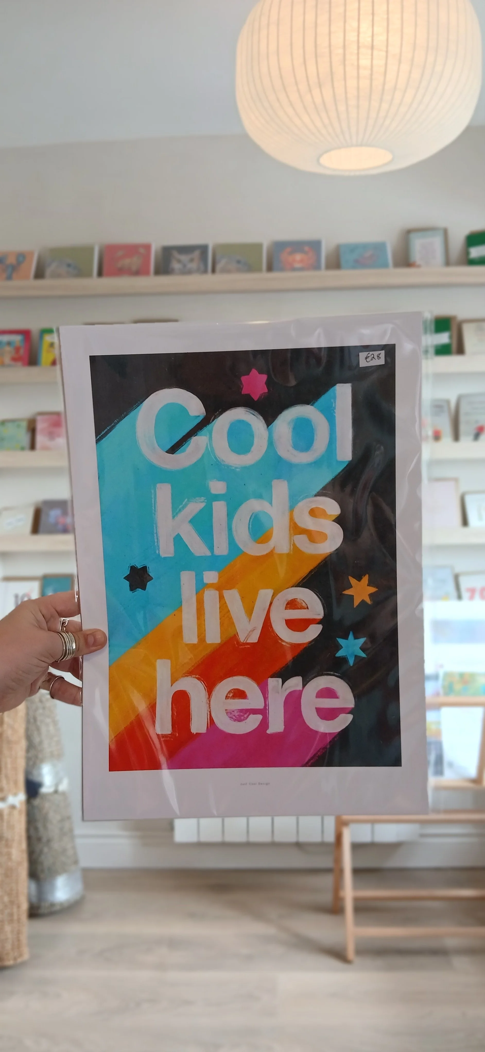 Colorful sign that says 'Cool kids live here' with stars and rainbow stripes, held in a store with bookshelves in the background.