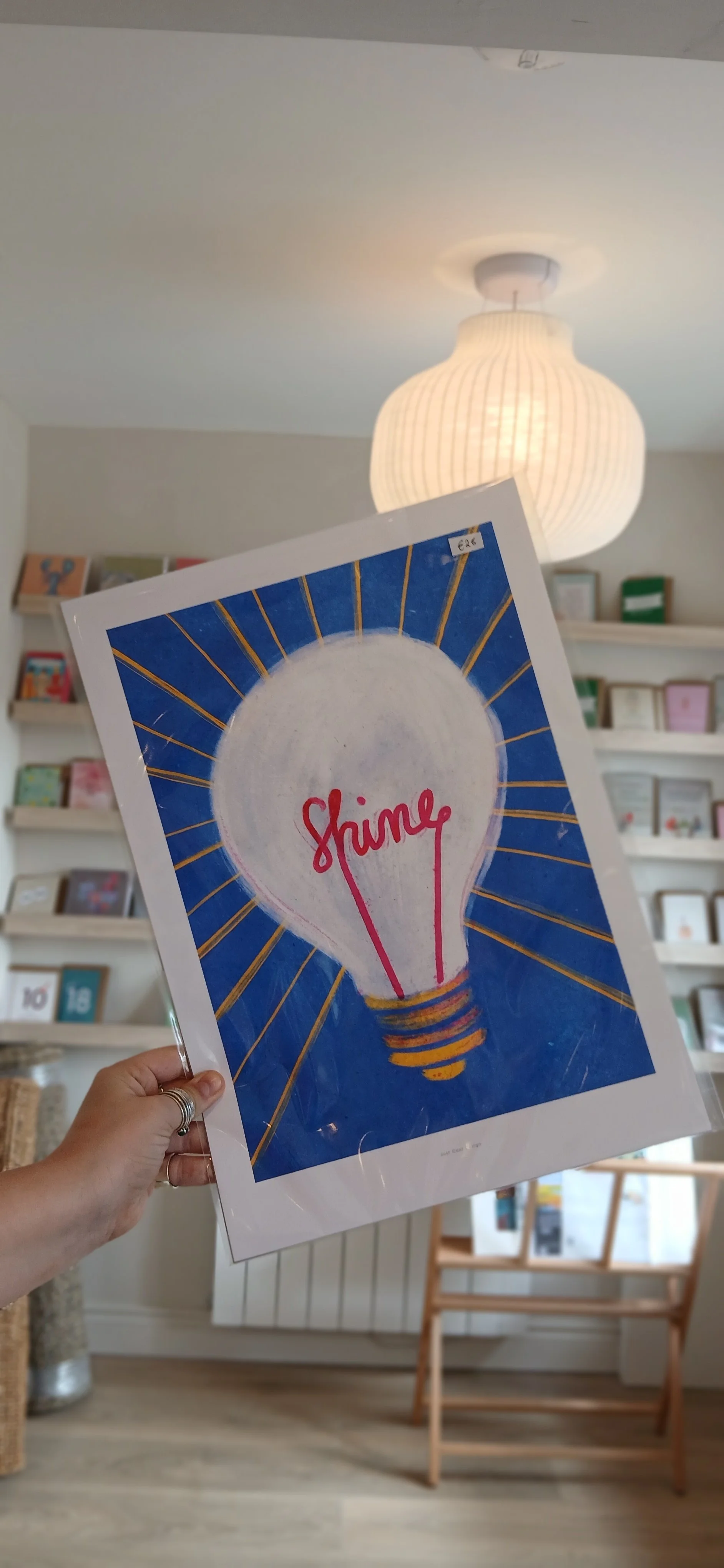 Hand holding a framed sign with a drawing of a light bulb and the word 'Shine' written inside. Background features a white bookshelf and a paper lantern ceiling light.