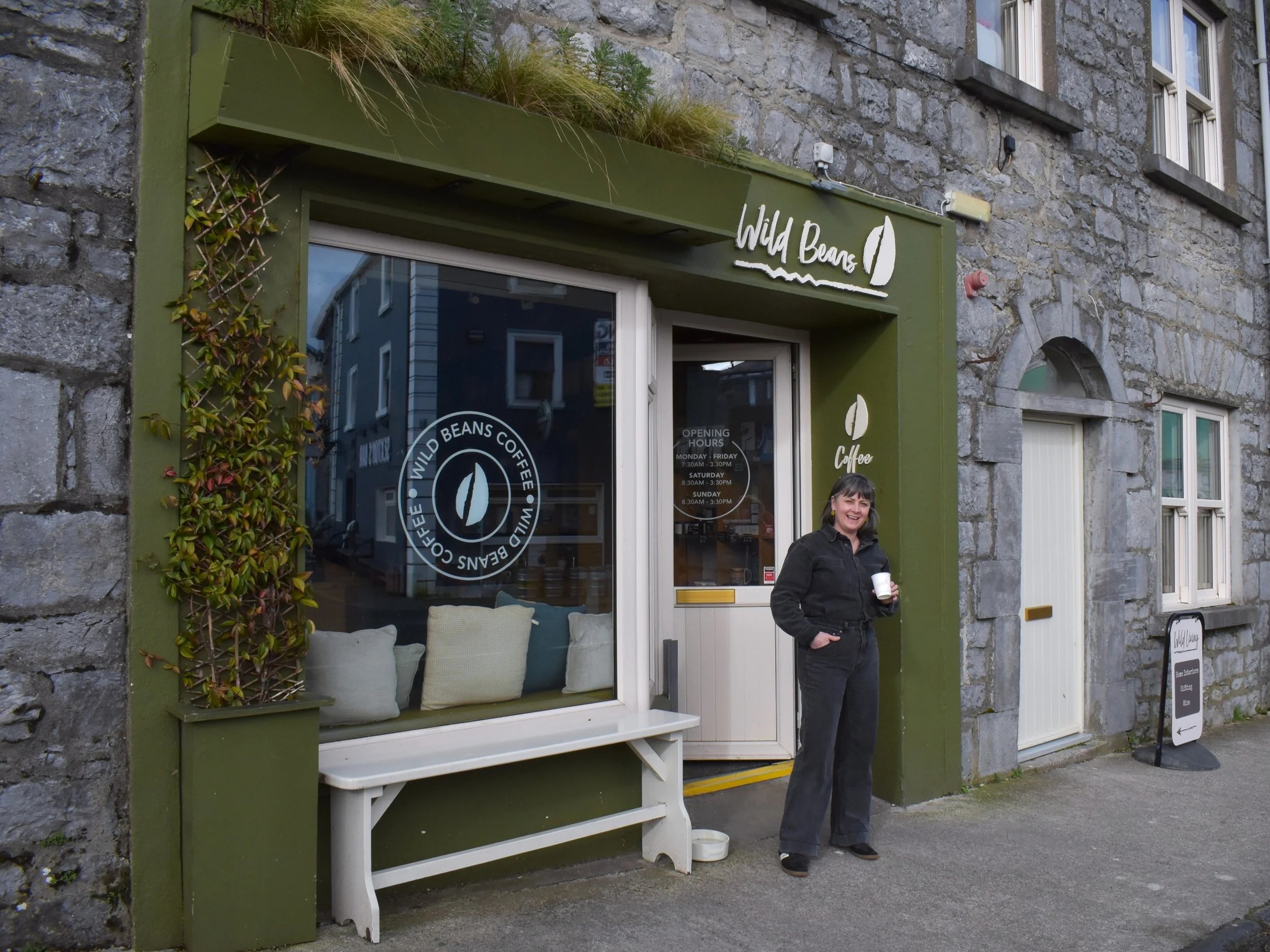 A woman standing outside a coffee shop called Wild Beans, holding a coffee cup and smiling. The shop has a green exterior with cushions in the window, and the sign says Wild Beans Coffee.