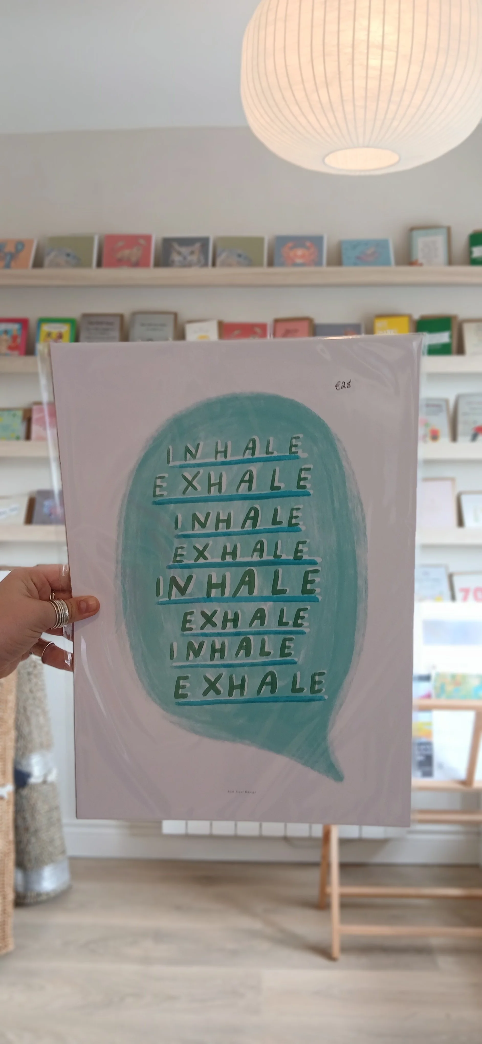A person holds a poster with the words "inhale" and "exhale" repeated in lines inside a speech bubble design with a bookstore or library in the background.
