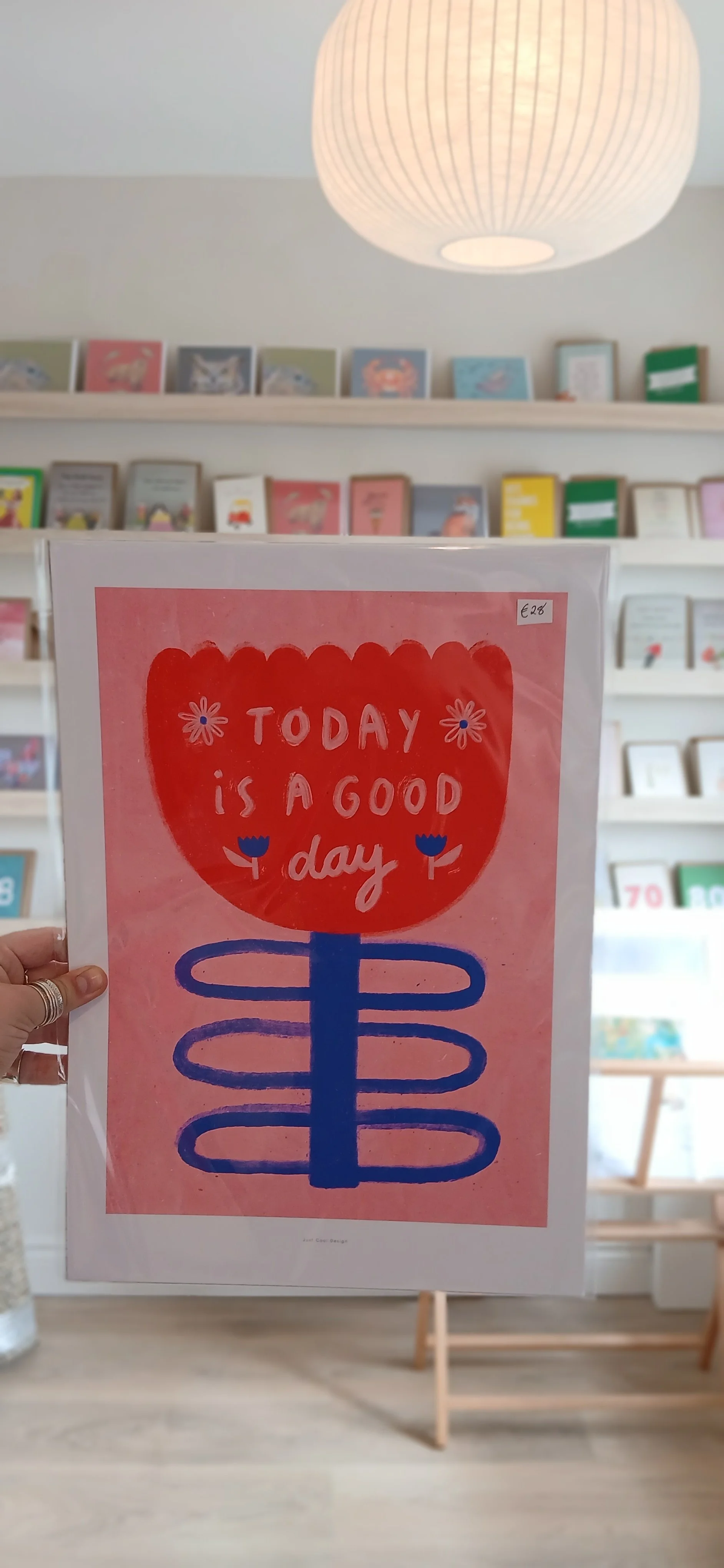 A colorful poster with a pink background and a red flower shape at the top, containing the words 'Today is a good day'. Below is a stylized blue swirl design. The poster is in a store with a bookshelf in the background and a large round ceiling light