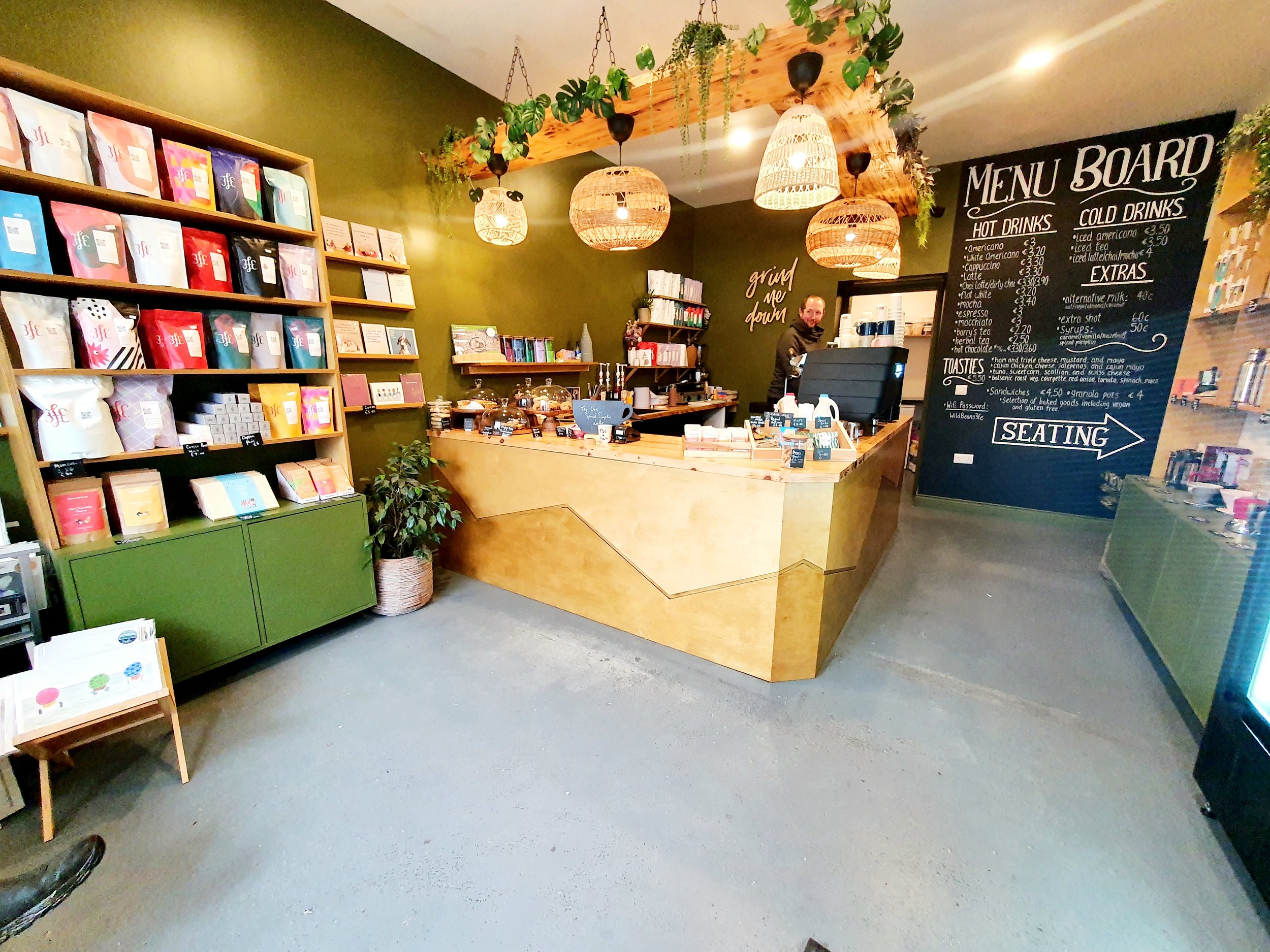 Interior of a cozy coffee shop with a green accent wall, wooden shelves with colorful packaged products, and a chalkboard menu listing hot and cold drinks. A counter with a barista serving, hanging wicker light fixtures, and potted plants add to the warm decor.