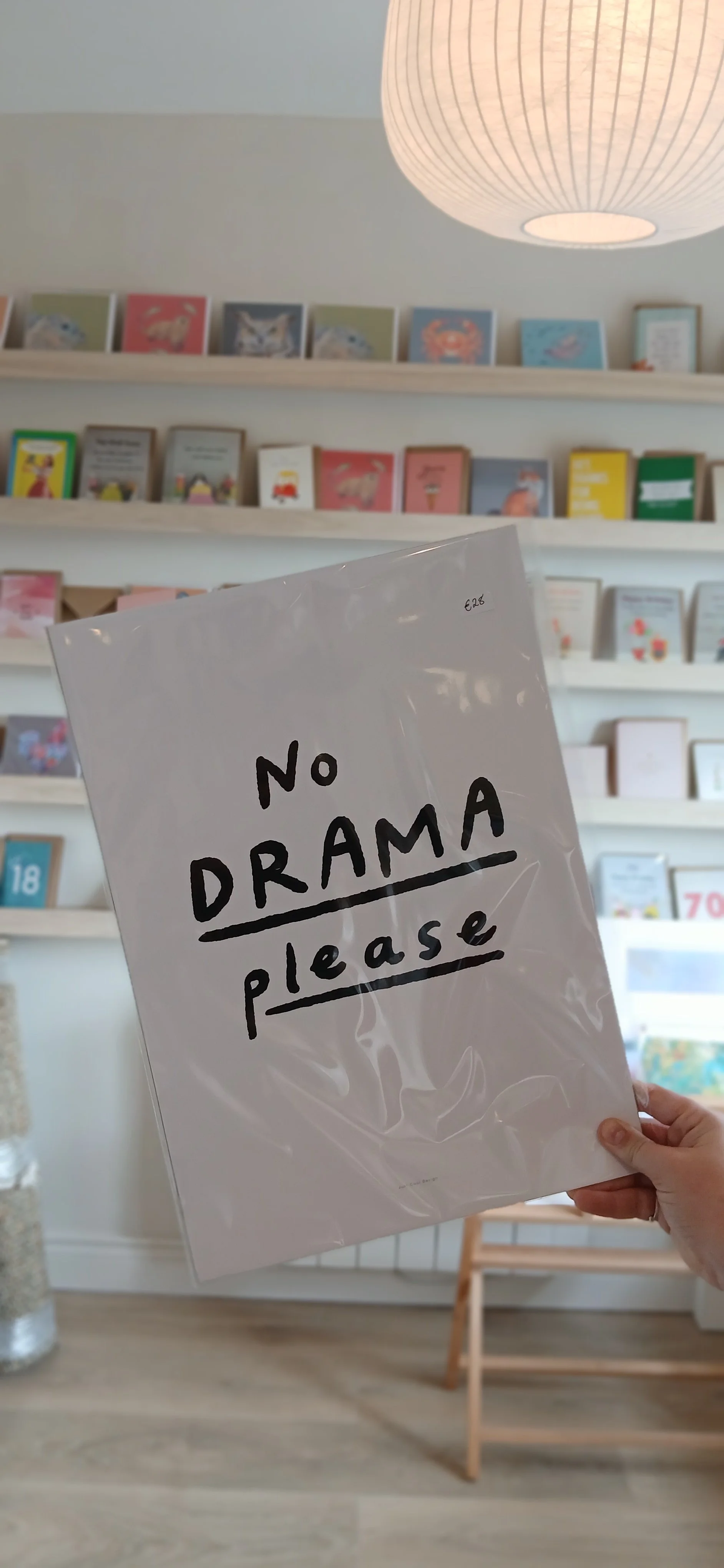 A person holding a framed print with the words 'No Drama Please' written in black letters, inside a store with shelves of books or cards in the background and a round ceiling light.