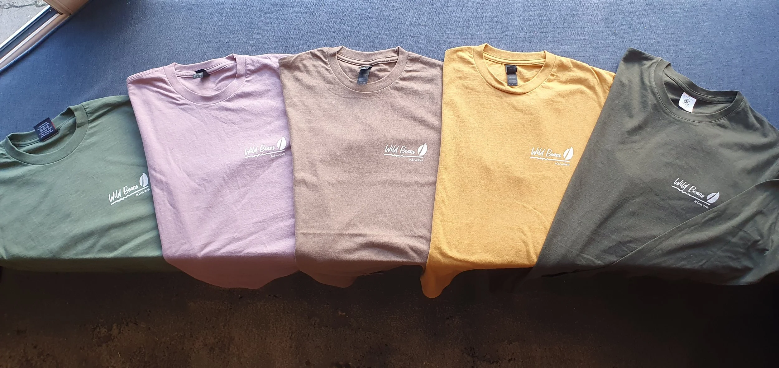 Five folded t-shirts in green, pink, beige, yellow, and dark green, each with a white logo that reads 'Wild Beans' on the front, laid out on a dark surface.