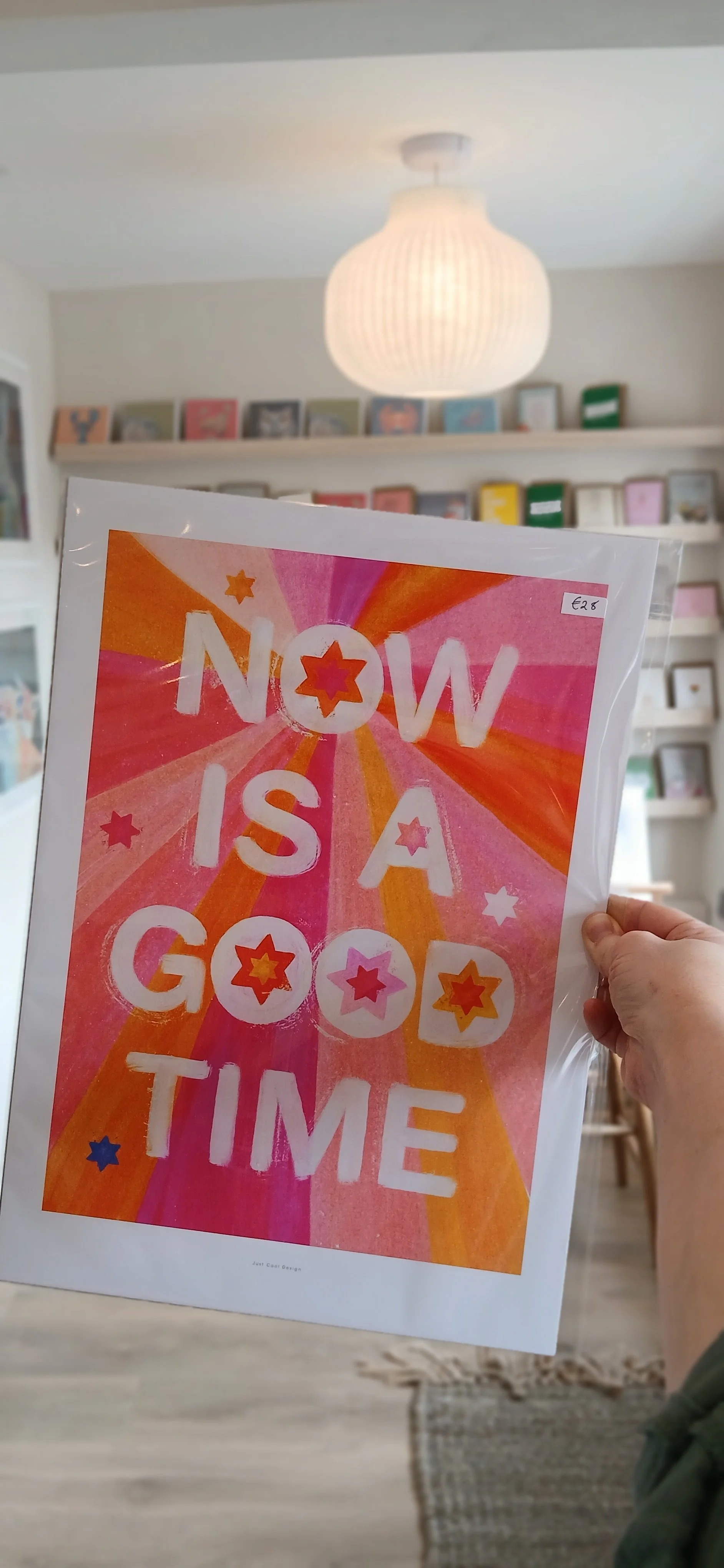 Person holding a colorful poster that says "Now is a good time" with star and burst graphics, in a room with shelves of books and framed pictures and a ceiling light.