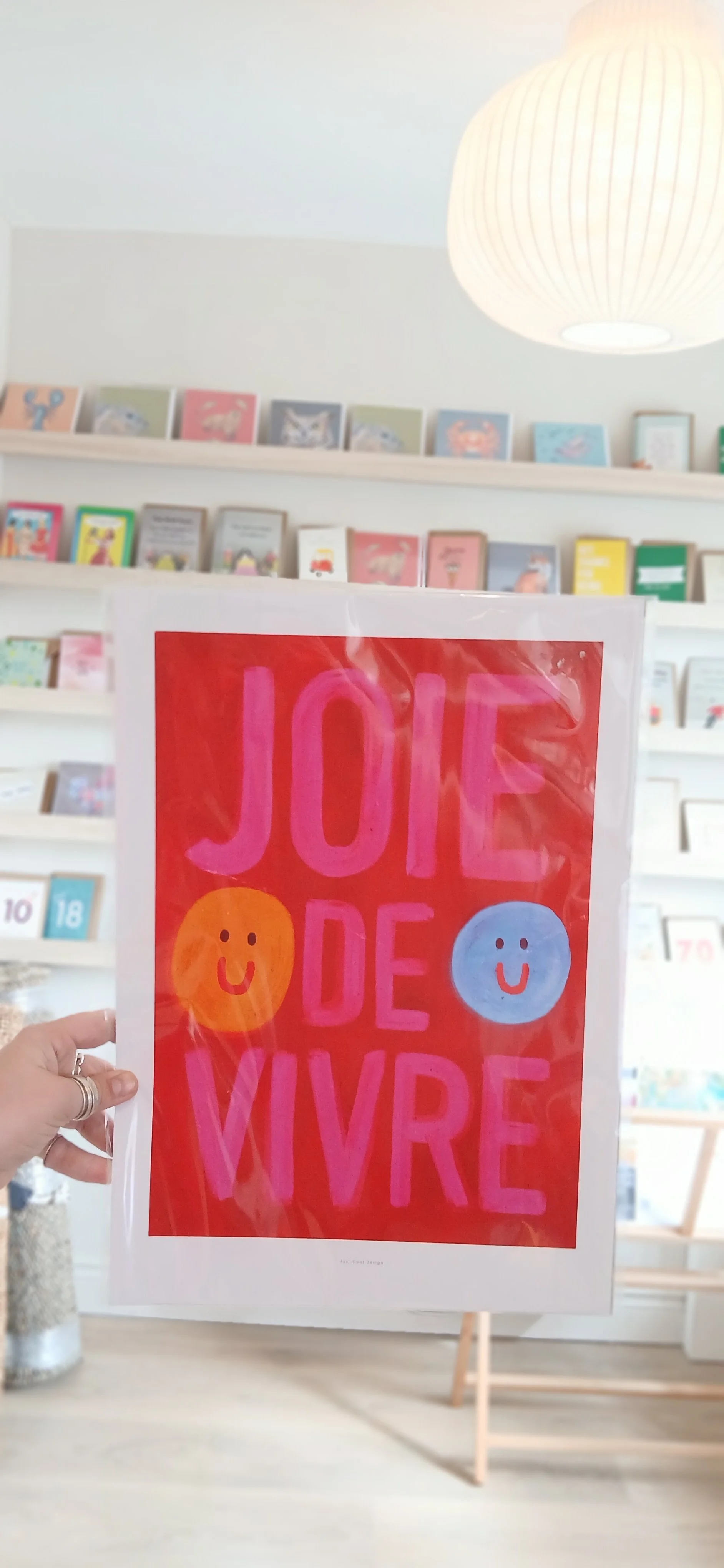A person holding a colorful poster that reads 'Joye de vivre' with smiley faces, inside a bookstore or library with shelves filled with books in the background.