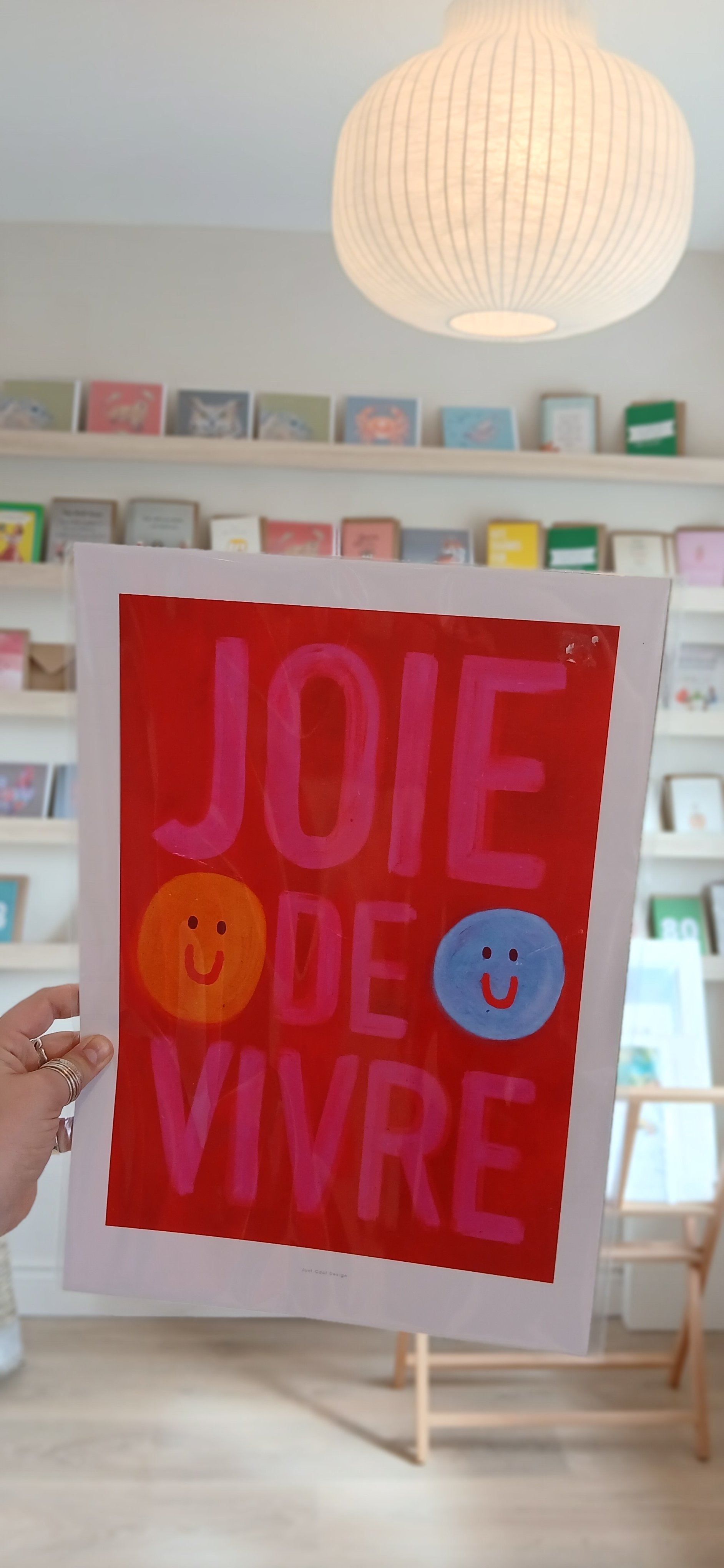 Hand holding a colorful poster with the words 'JOIE DE VIVRE' and smiley faces in a bright, well-lit room with bookshelves and a hanging paper lantern.